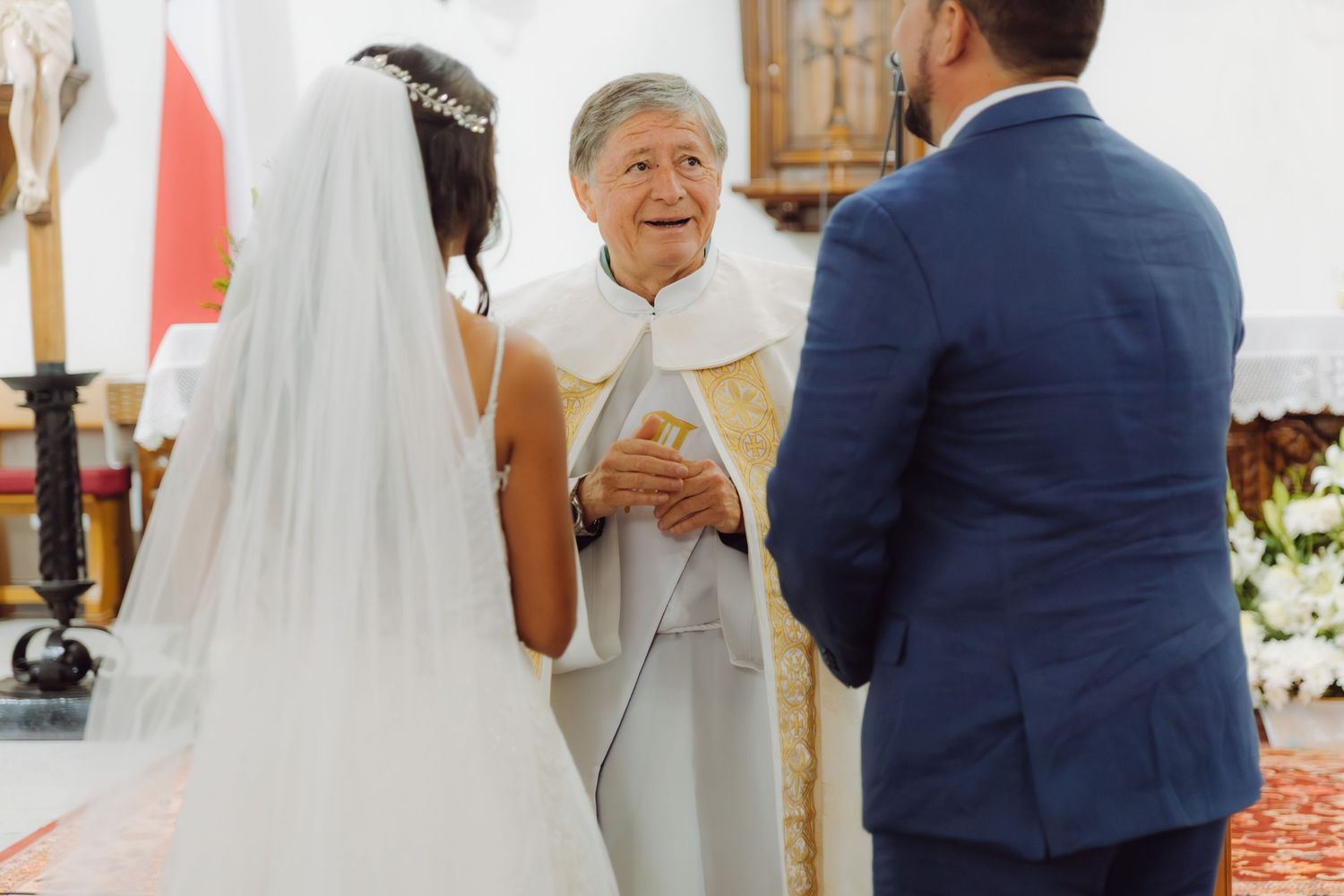 Smiling elderly priest officiates wedding ceremony with bride in tiara veil and groom in blue suit.