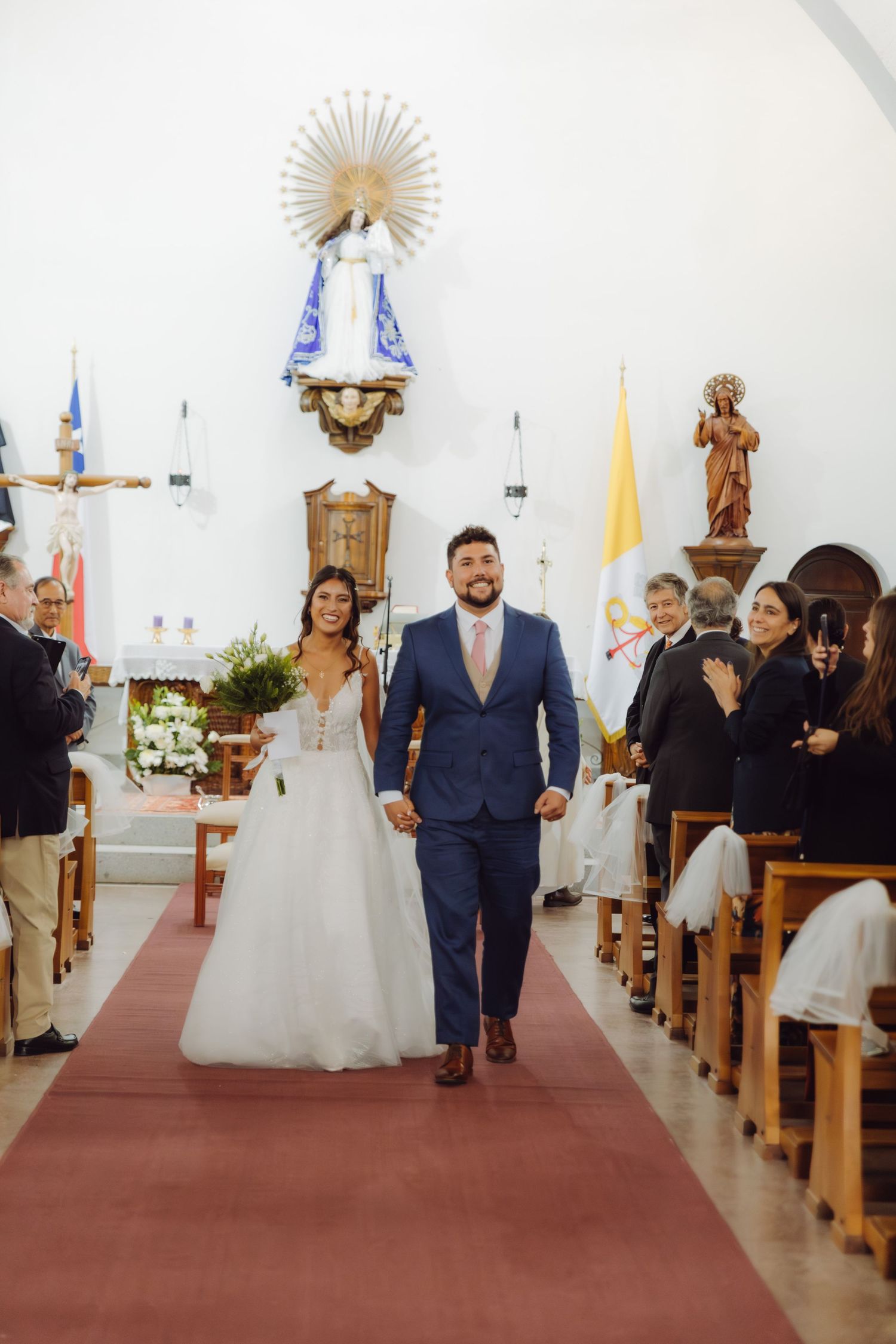 Joyful newlyweds walk down red carpet aisle after wedding ceremony in decorated Catholic church.