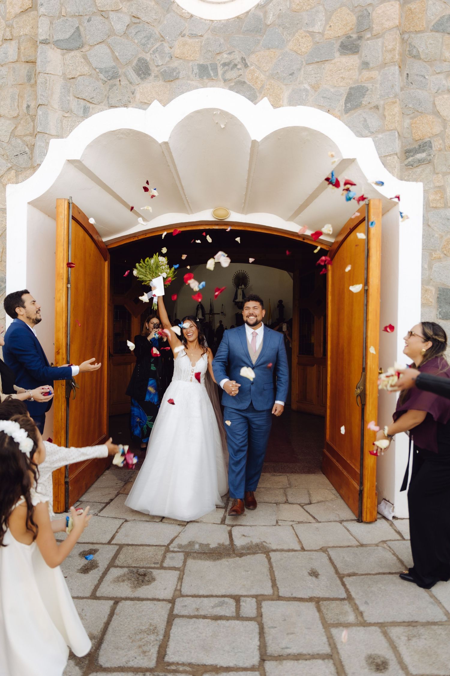 Newlyweds exit church through arched wooden doors as guests throw colorful confetti petals.