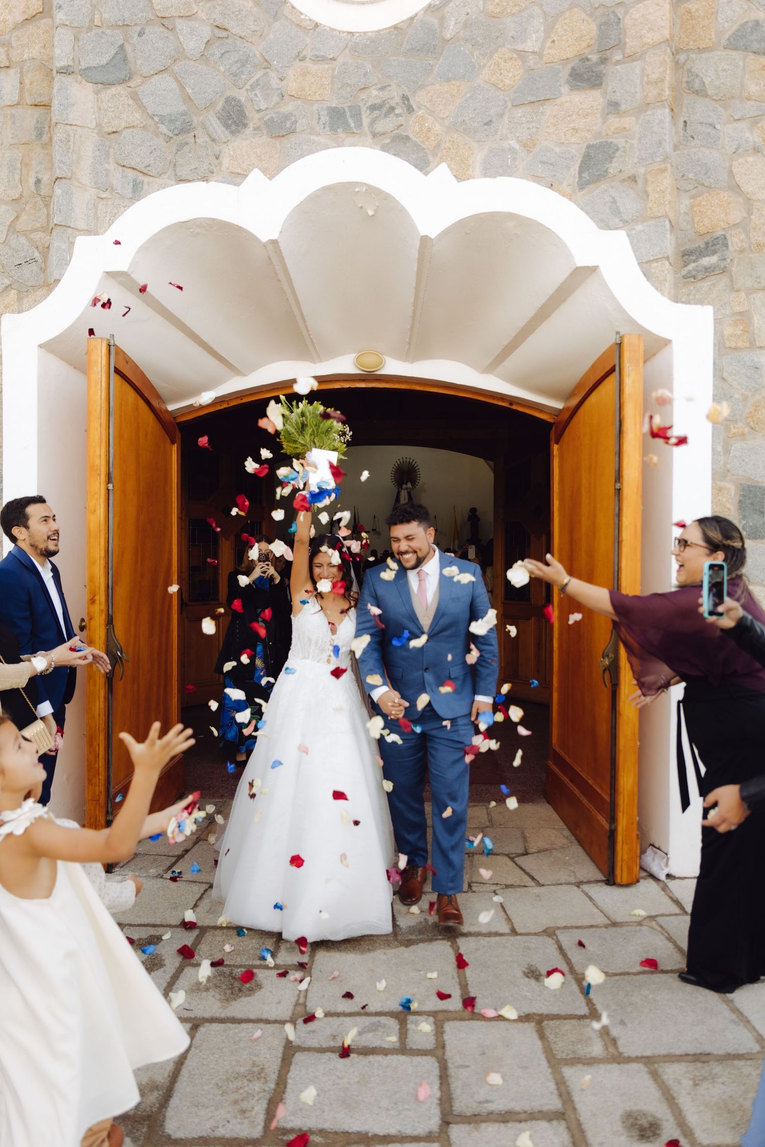 Newlywed couple exits church doors as guests toss colorful flower petals in joyful wedding celebration.
