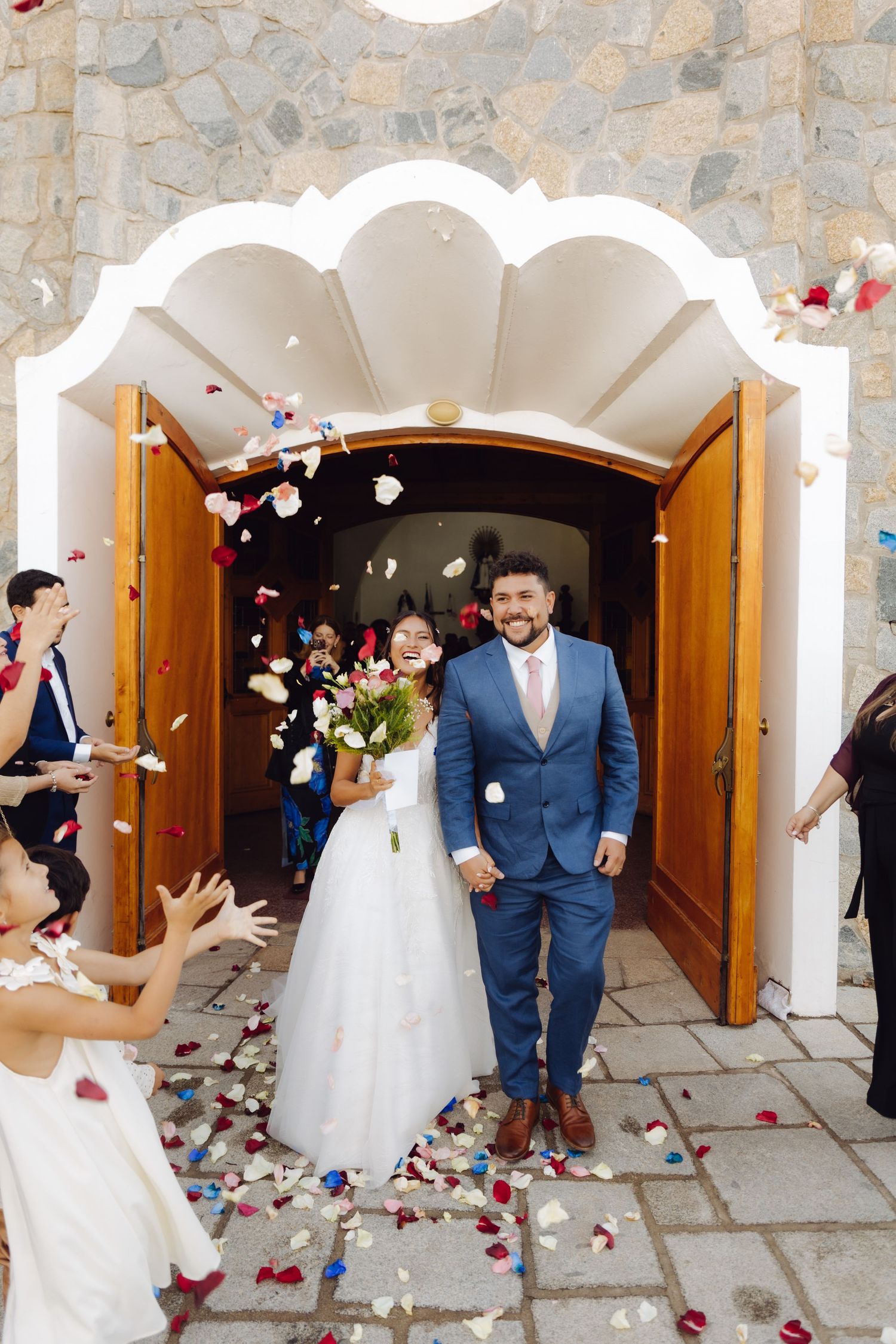 Bride and groom walk out of church entrance surrounded by guests throwing red and white flower petals.