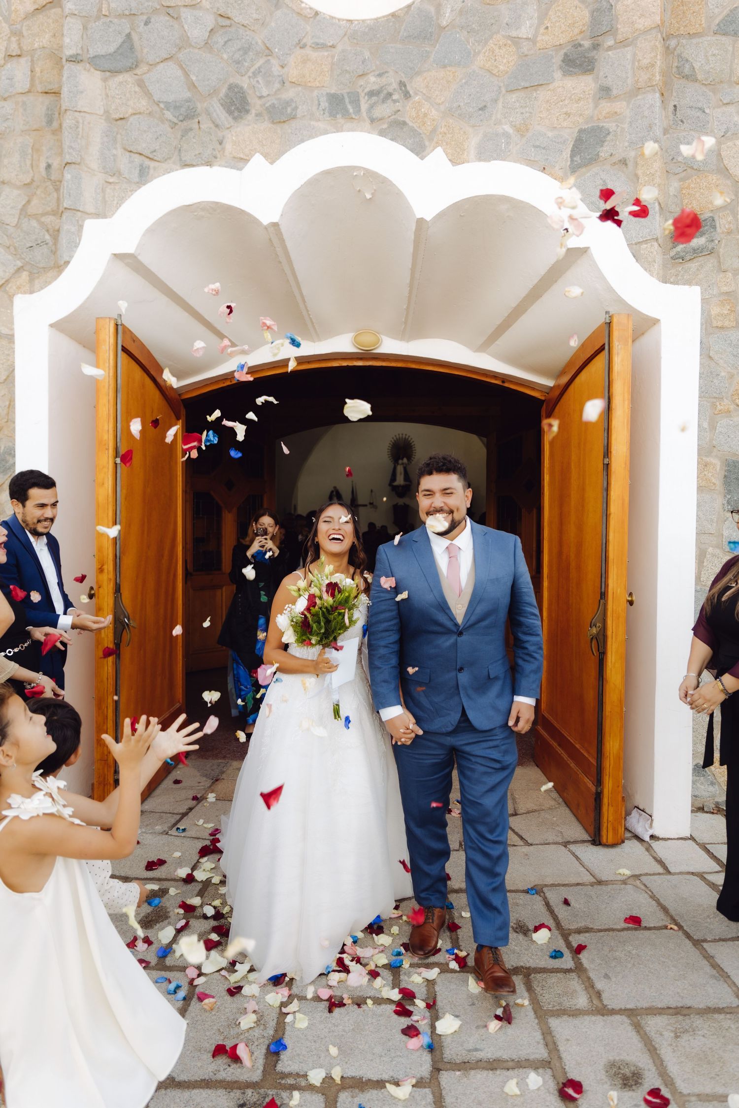 Happy newlyweds hold hands exiting church as colorful confetti petals fill the air during wedding.