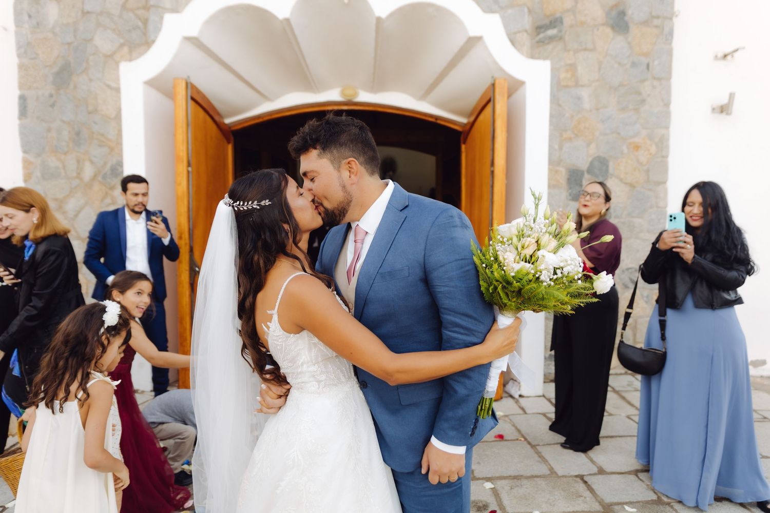 Bride and groom share a romantic kiss outside church doors surrounded by wedding guests and flowers.