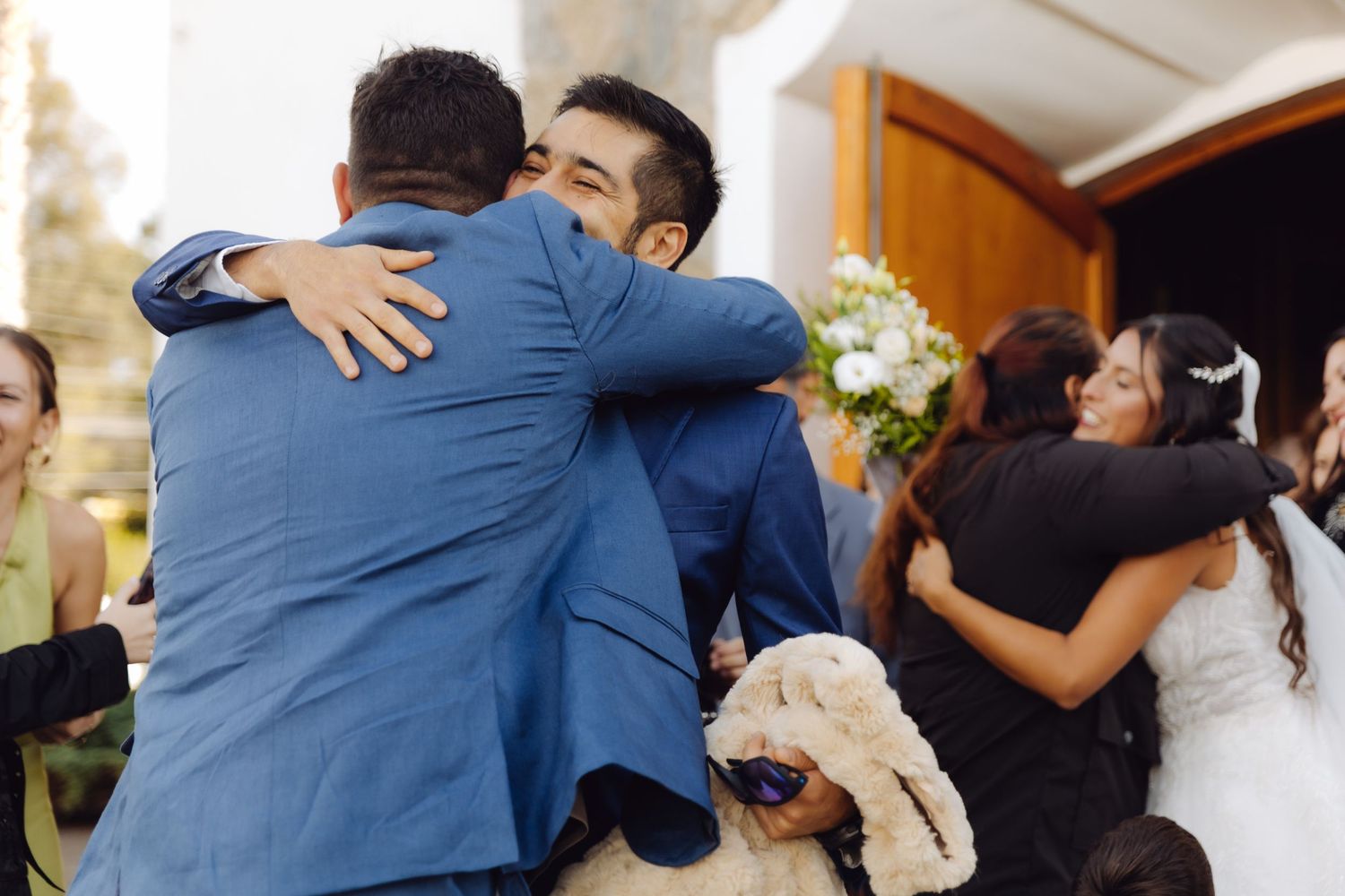 Groom in blue suit shares warm embrace with male guest while bride hugs another guest nearby.