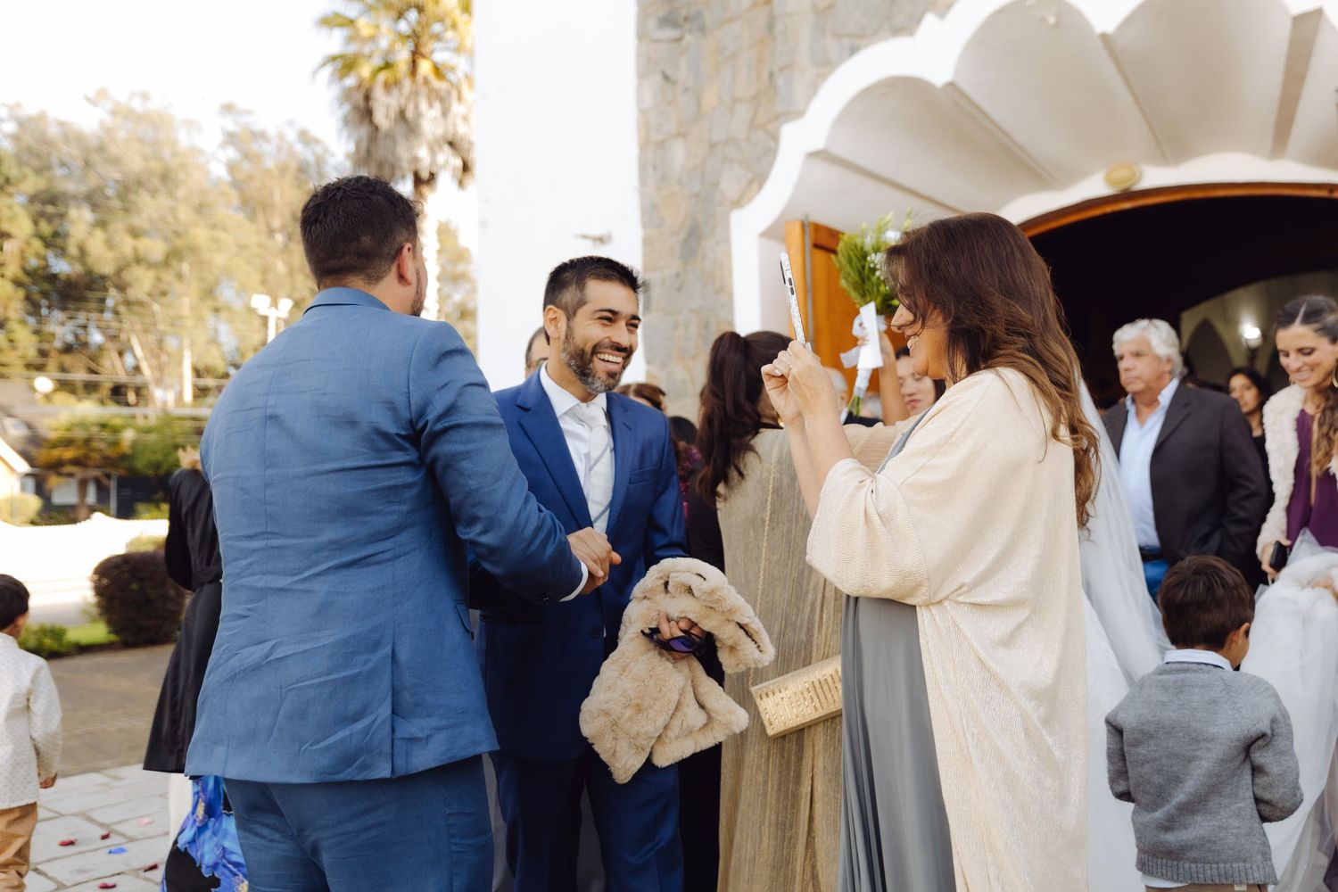 Wedding guests in formal attire greet and congratulate the groom outside a white church venue.