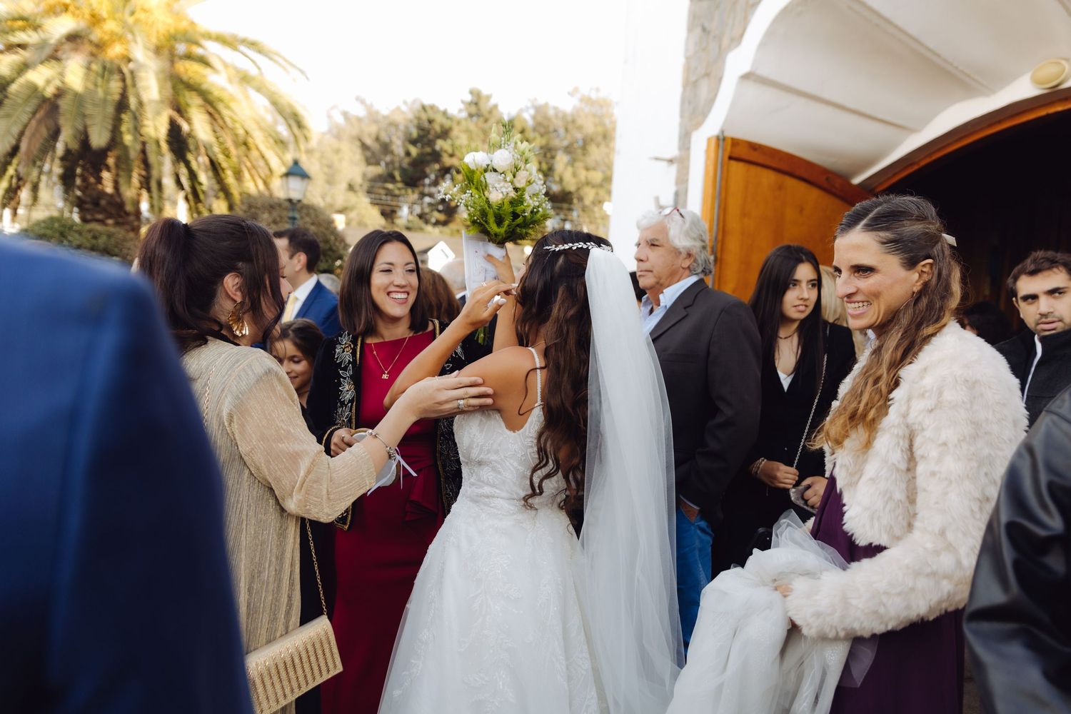 Bride in white wedding gown receives warm congratulations from guests outside church after ceremony.