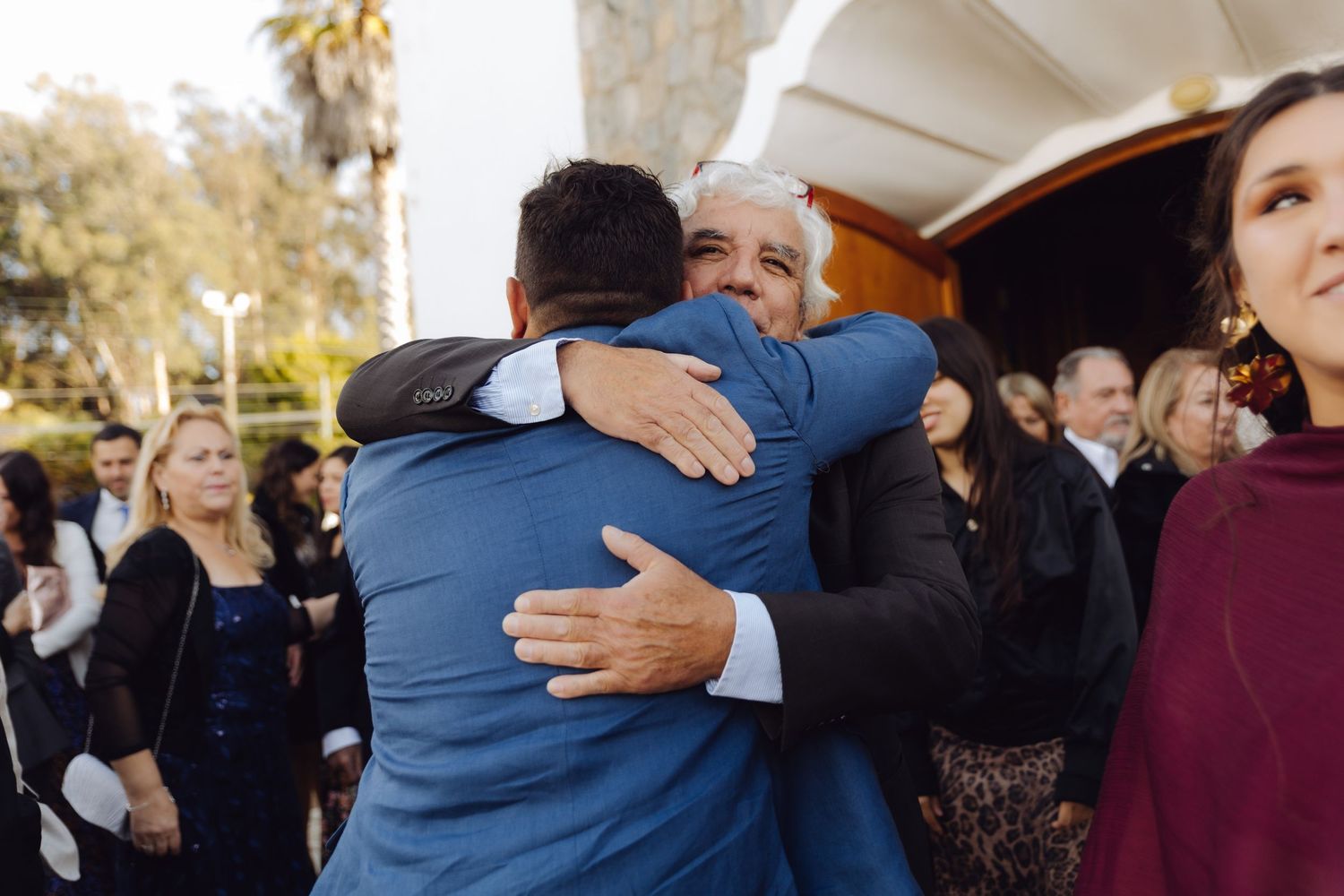 Groom in blue suit shares heartfelt embrace with older male guest outside church after wedding.