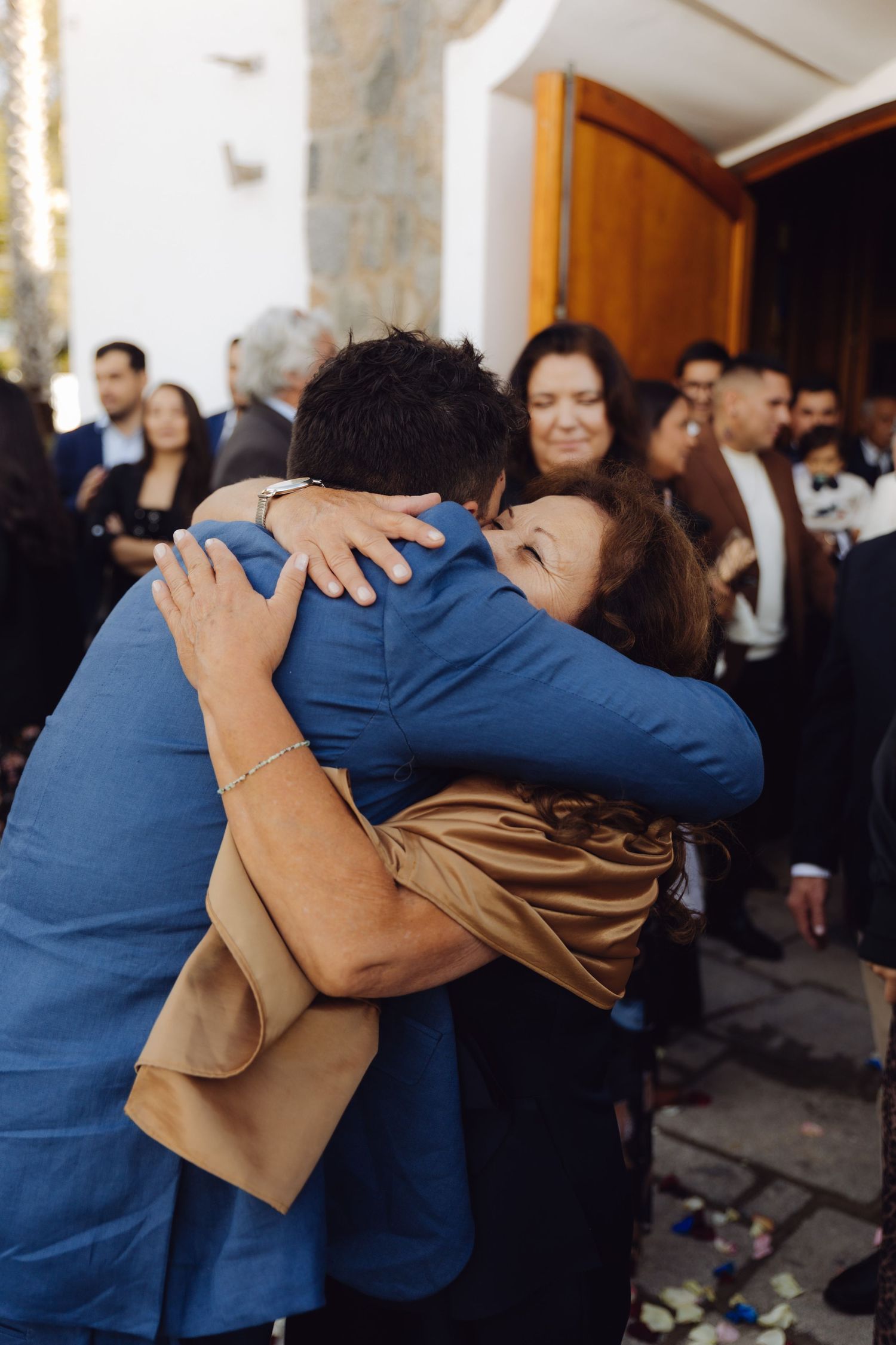 Groom in blue suit receives warm hug from older woman guest outside church after wedding ceremony.