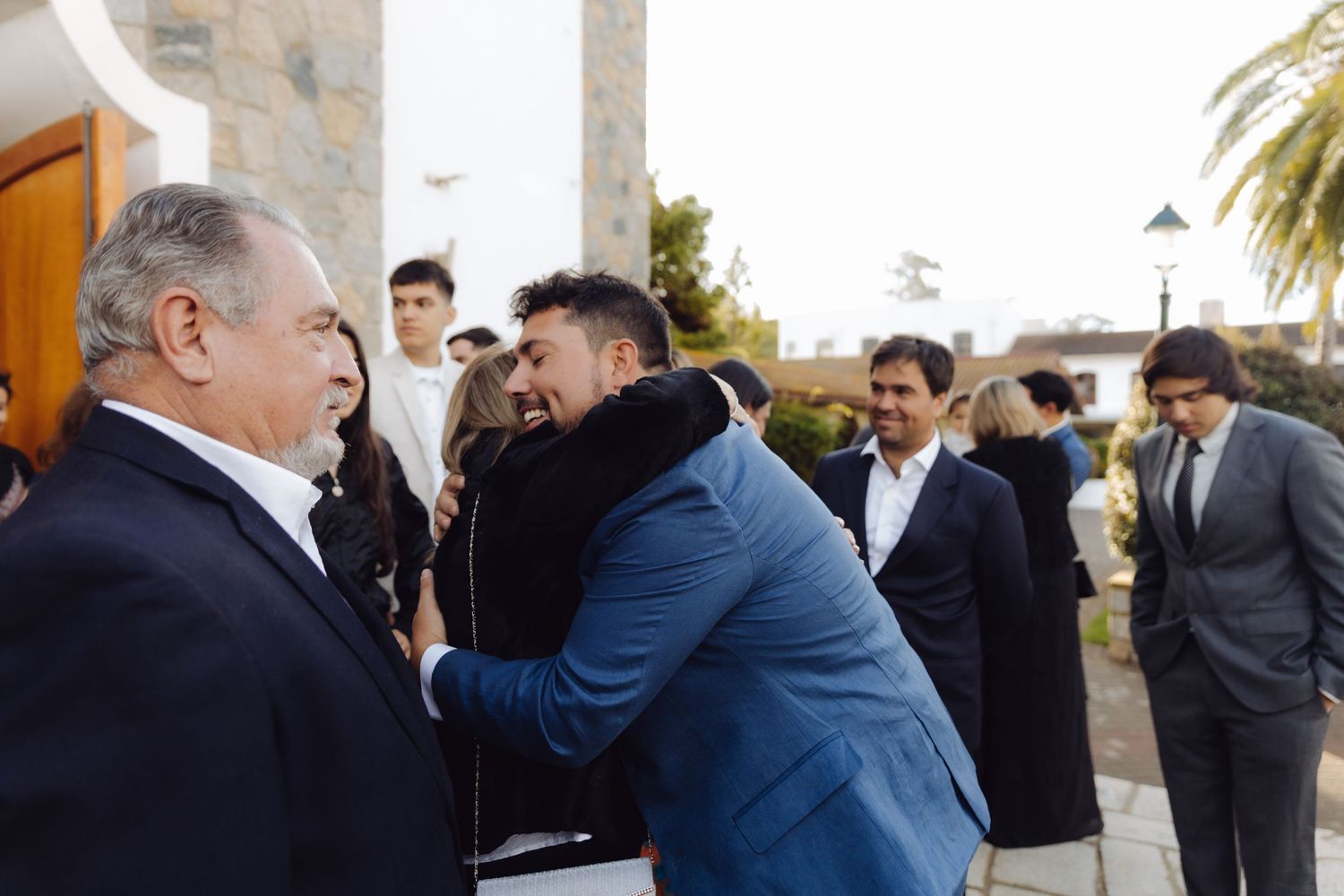 Wedding guests embracing and laughing outside a white chapel during a reception.