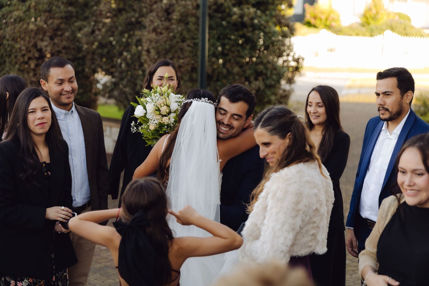 Bride and groom sharing a hug surrounded by smiling wedding guests outdoors.