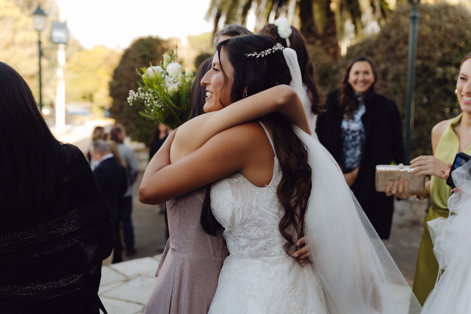 Bride in white lace wedding dress sharing a warm hug with a guest in a mauve dress.