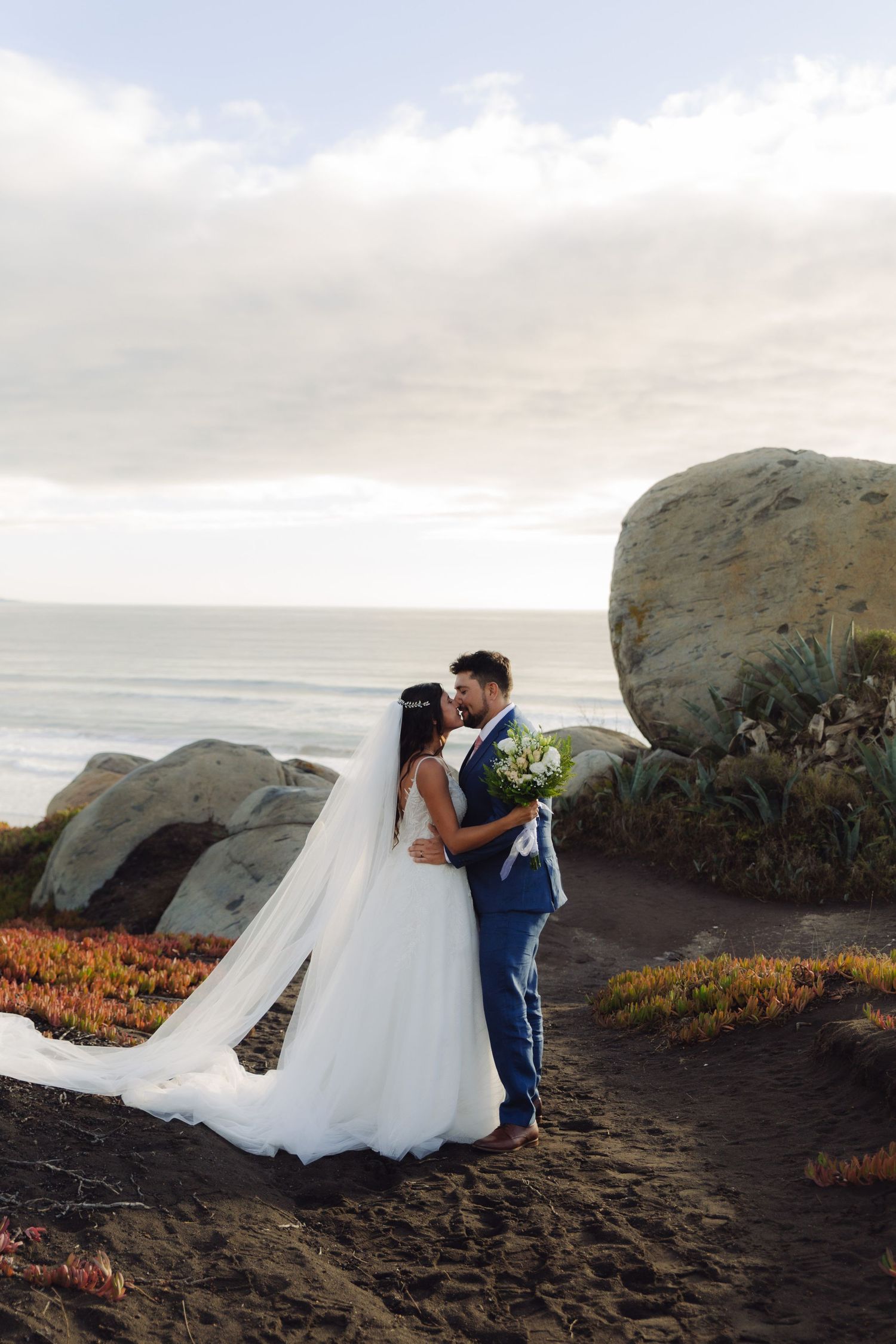 Bride and groom kissing near ocean rocks with a flowing veil and white bouquet.