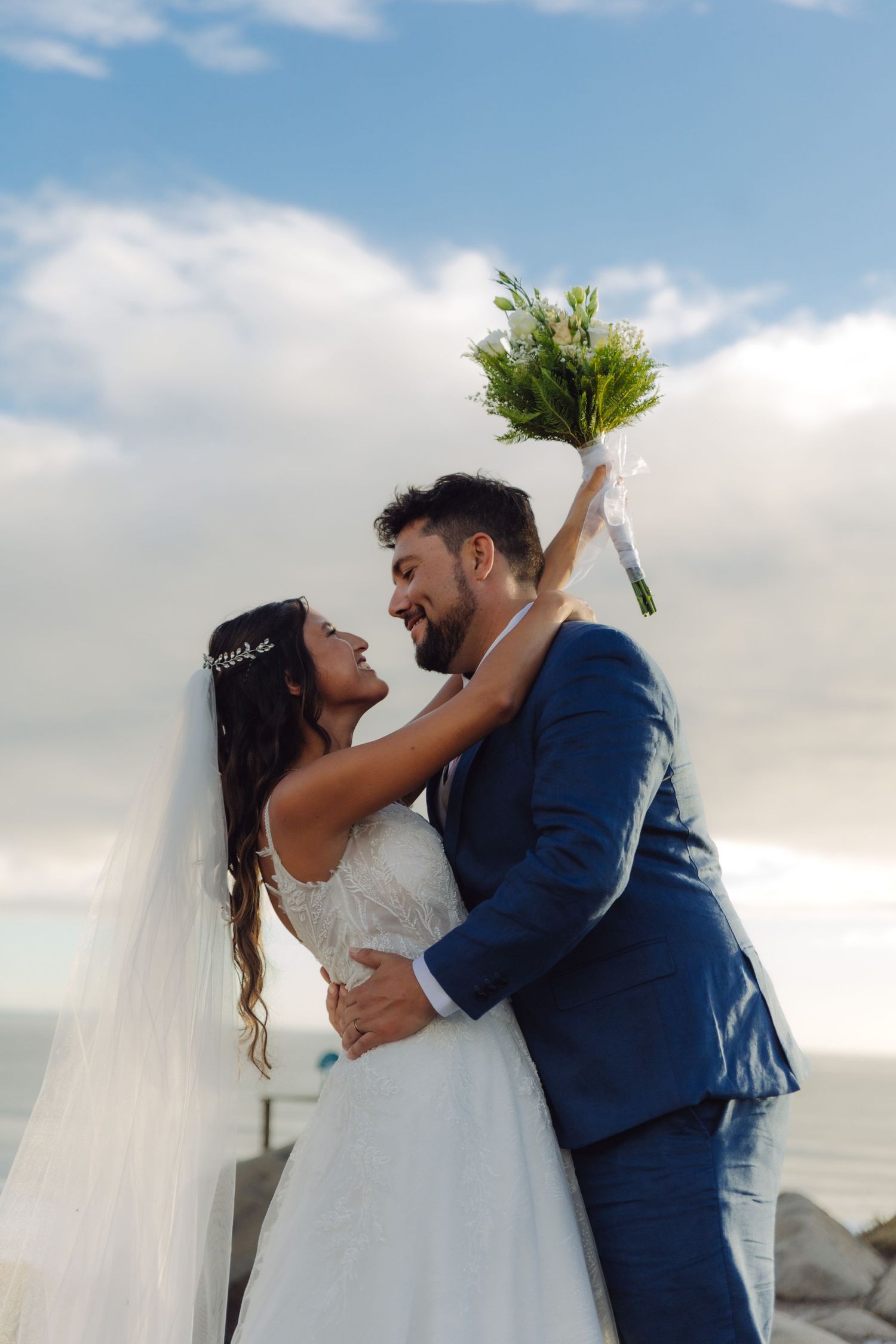 Joyful bride and groom embracing with bouquet raised against a blue cloudy sky.
