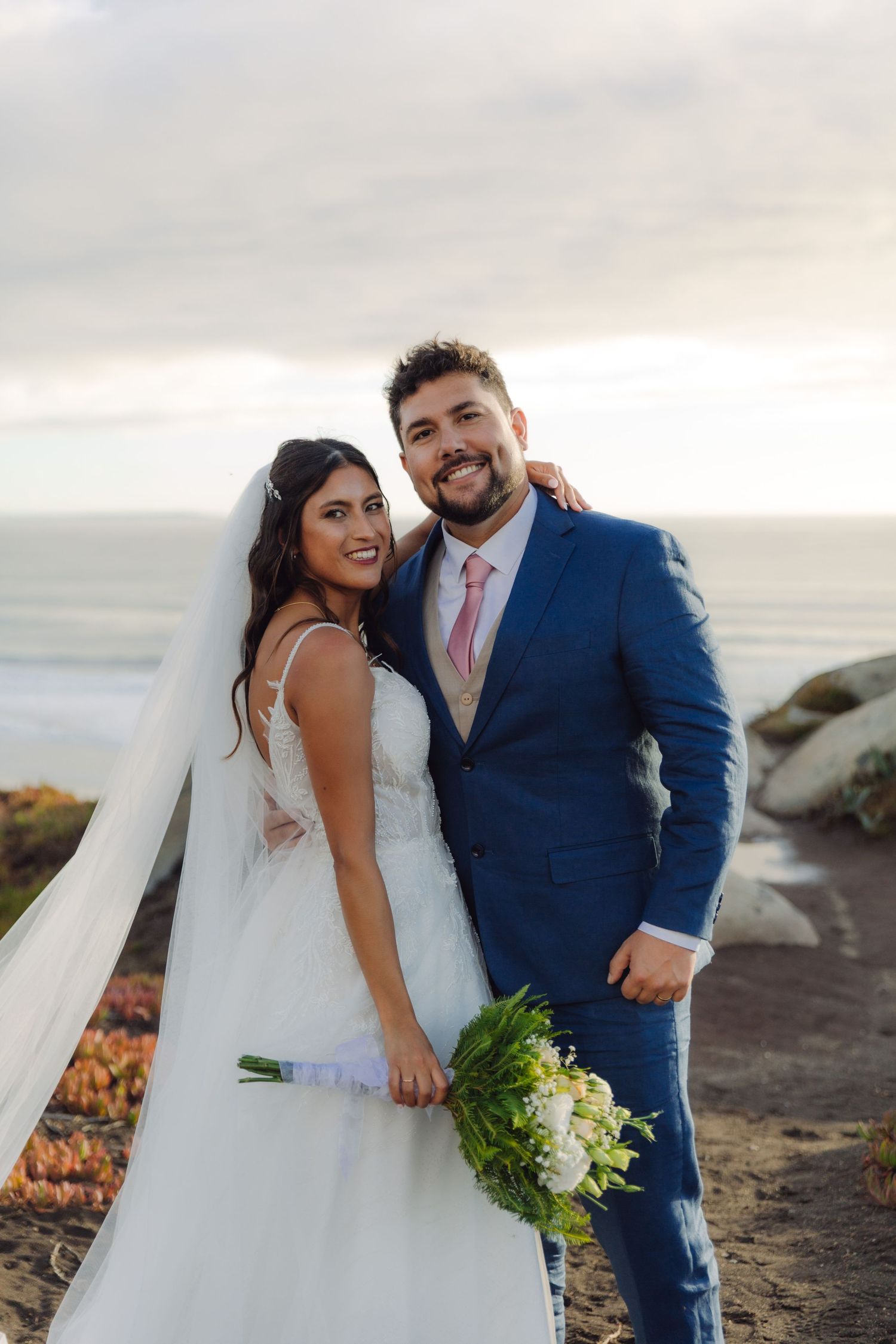 Smiling bride and groom posing together with white bouquet near a scenic ocean cliff.