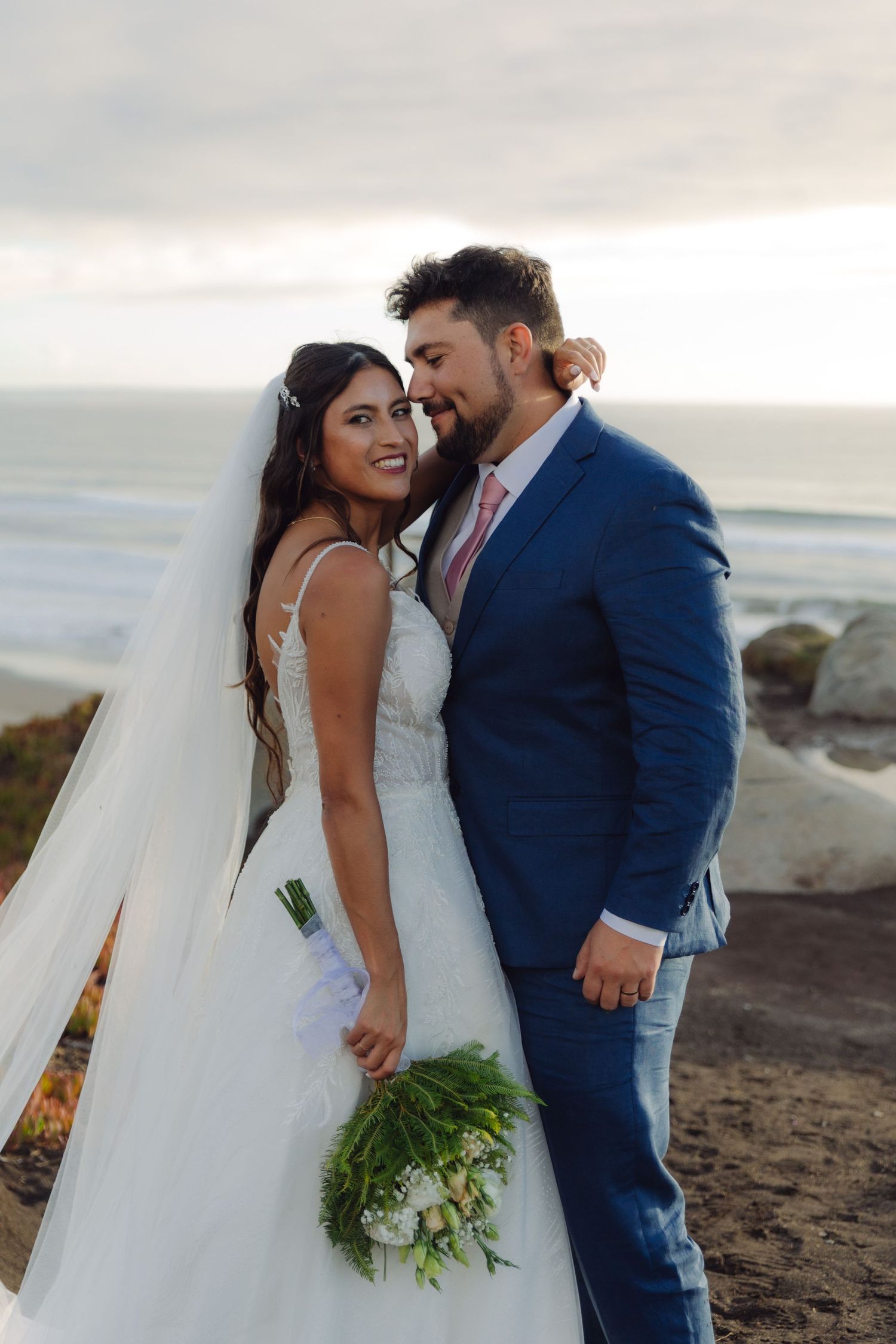 Bride and groom share a romantic moment on a coastal bluff, she in a white gown with veil, he in a blue suit.