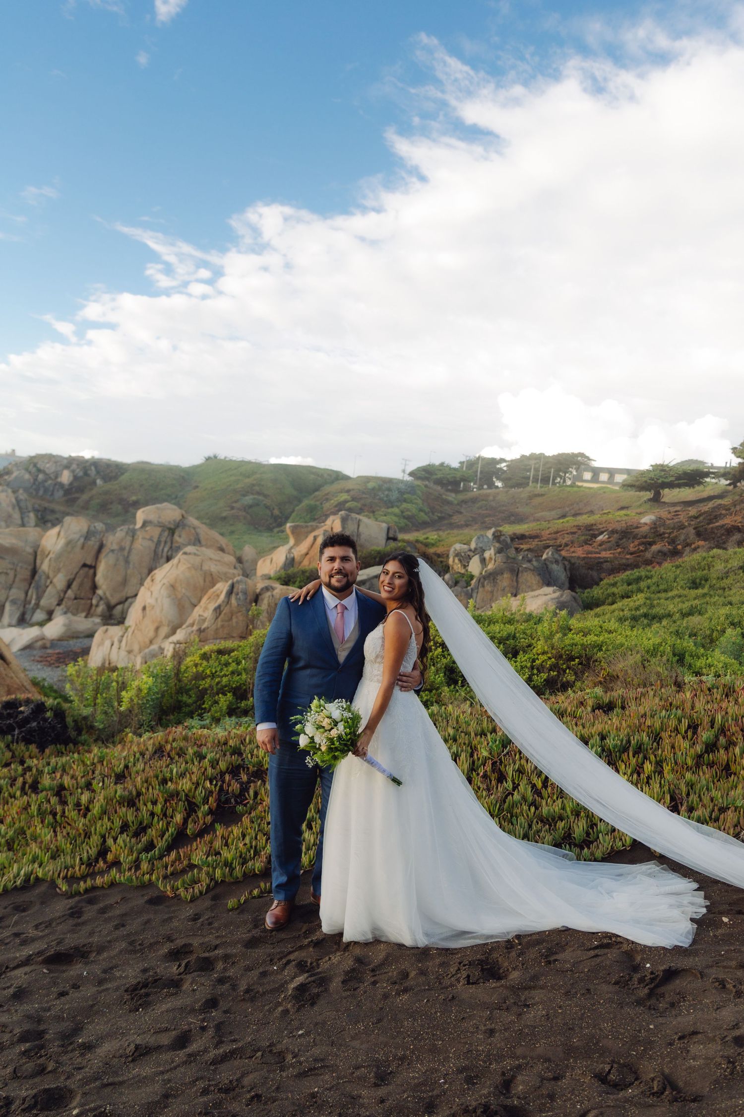 Wedding couple poses on a rocky beach, bride's long veil flowing in the wind with green hills behind them.