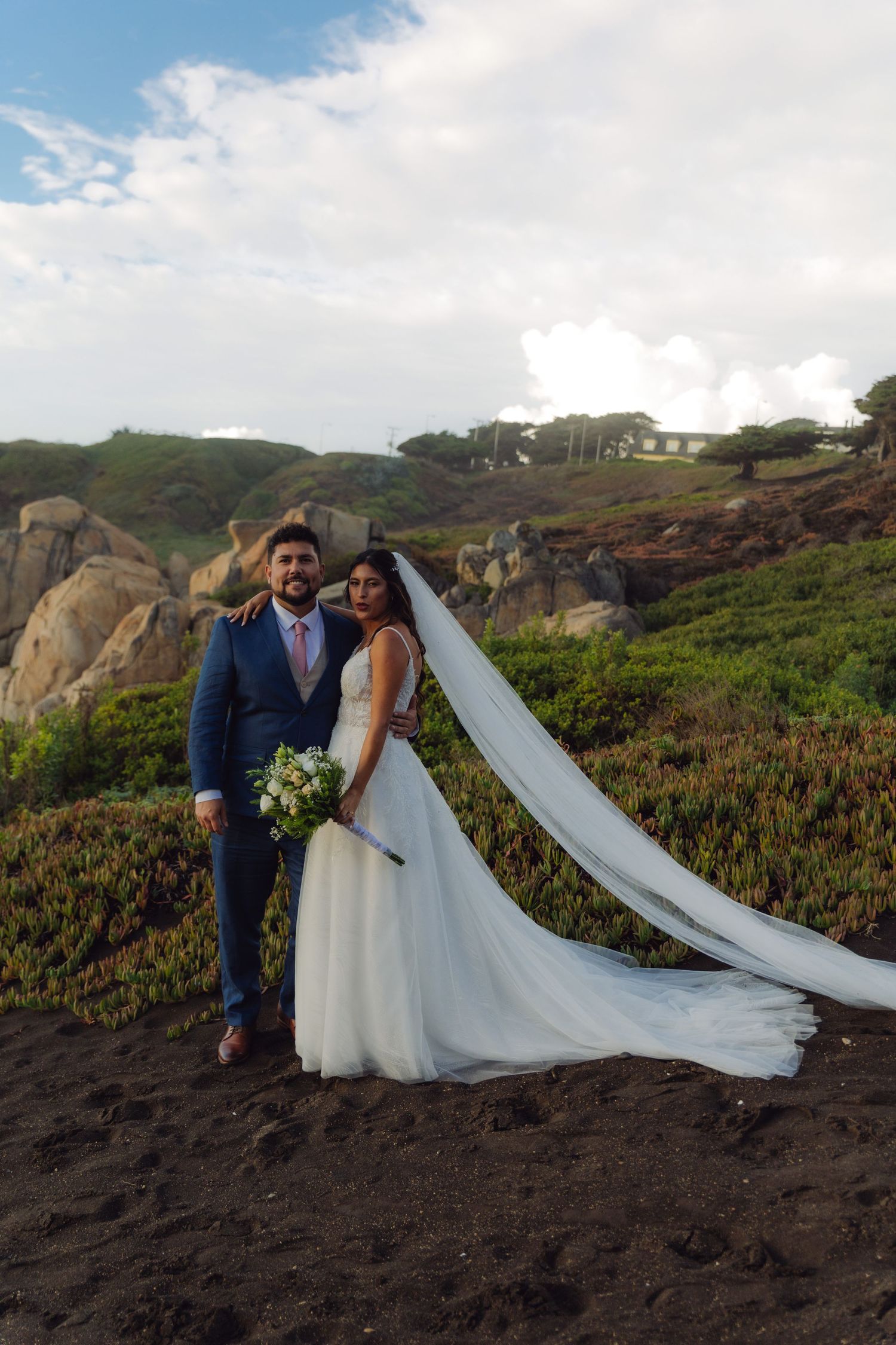 Newlyweds stand together on a dark sandy beach, bride holding bouquet with veil blowing, rocky cliffs behind.