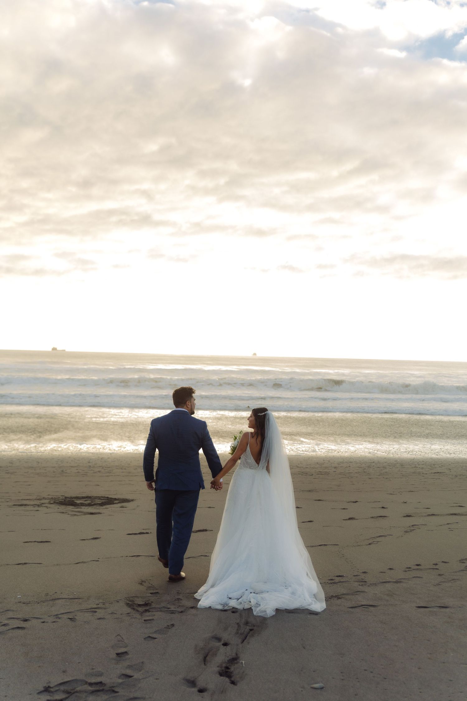 Bride and groom walk hand-in-hand toward the ocean at sunset, leaving footprints in the wet sand.