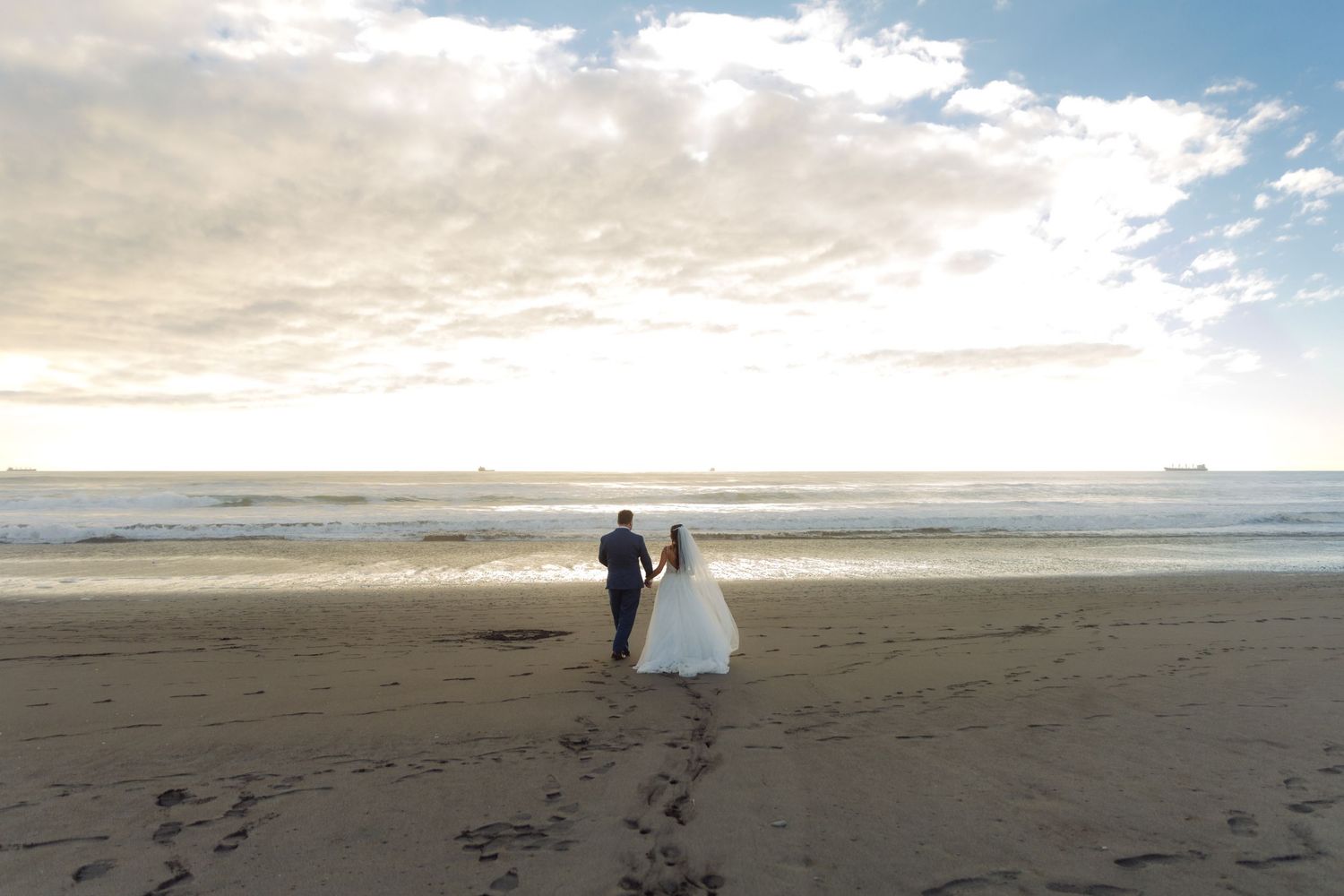 Distant view of bride and groom standing together on a wide sandy beach at sunset with ocean waves.