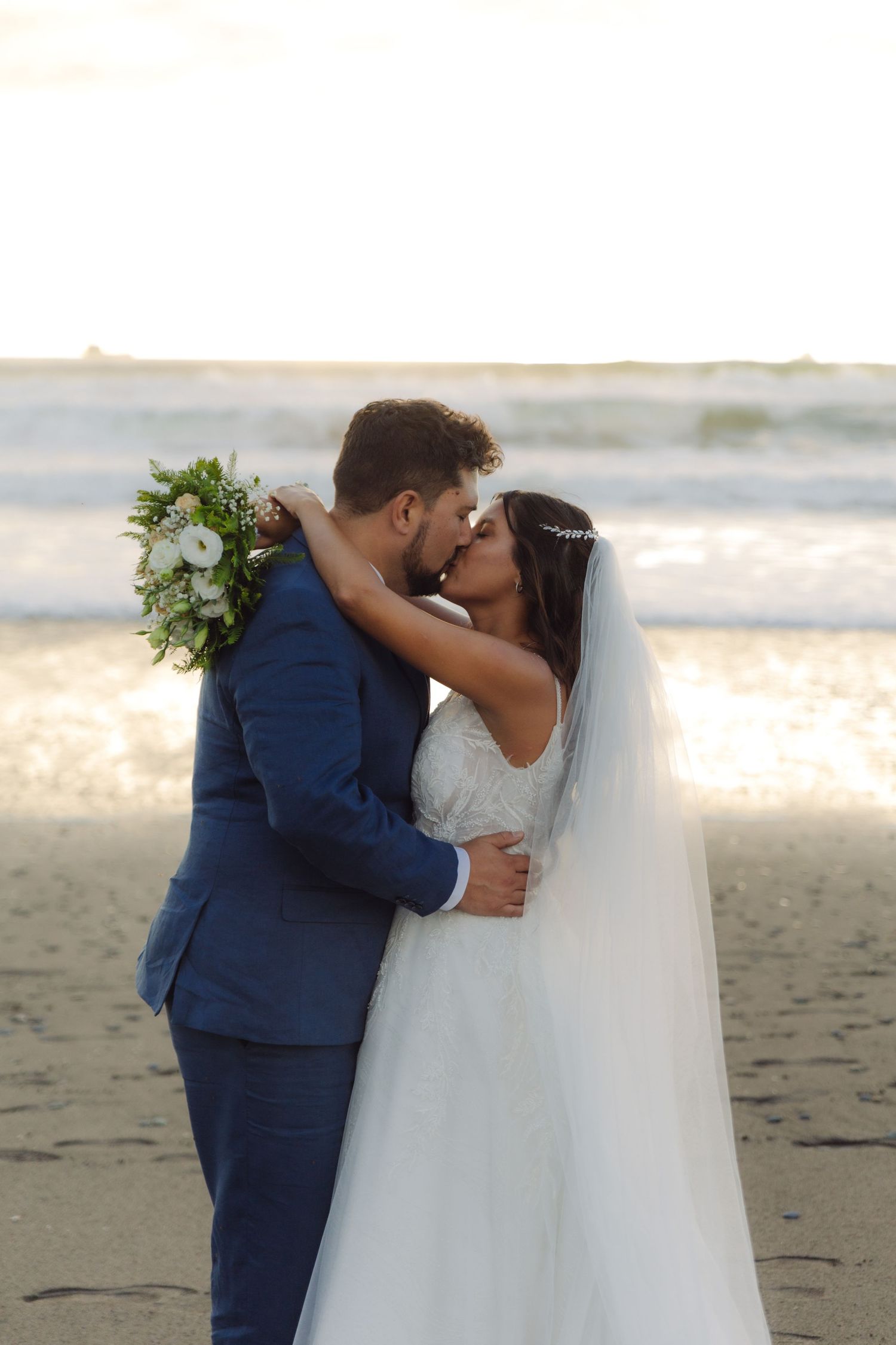 Bride and groom share a passionate kiss on the beach at golden hour, bride holding white floral bouquet.