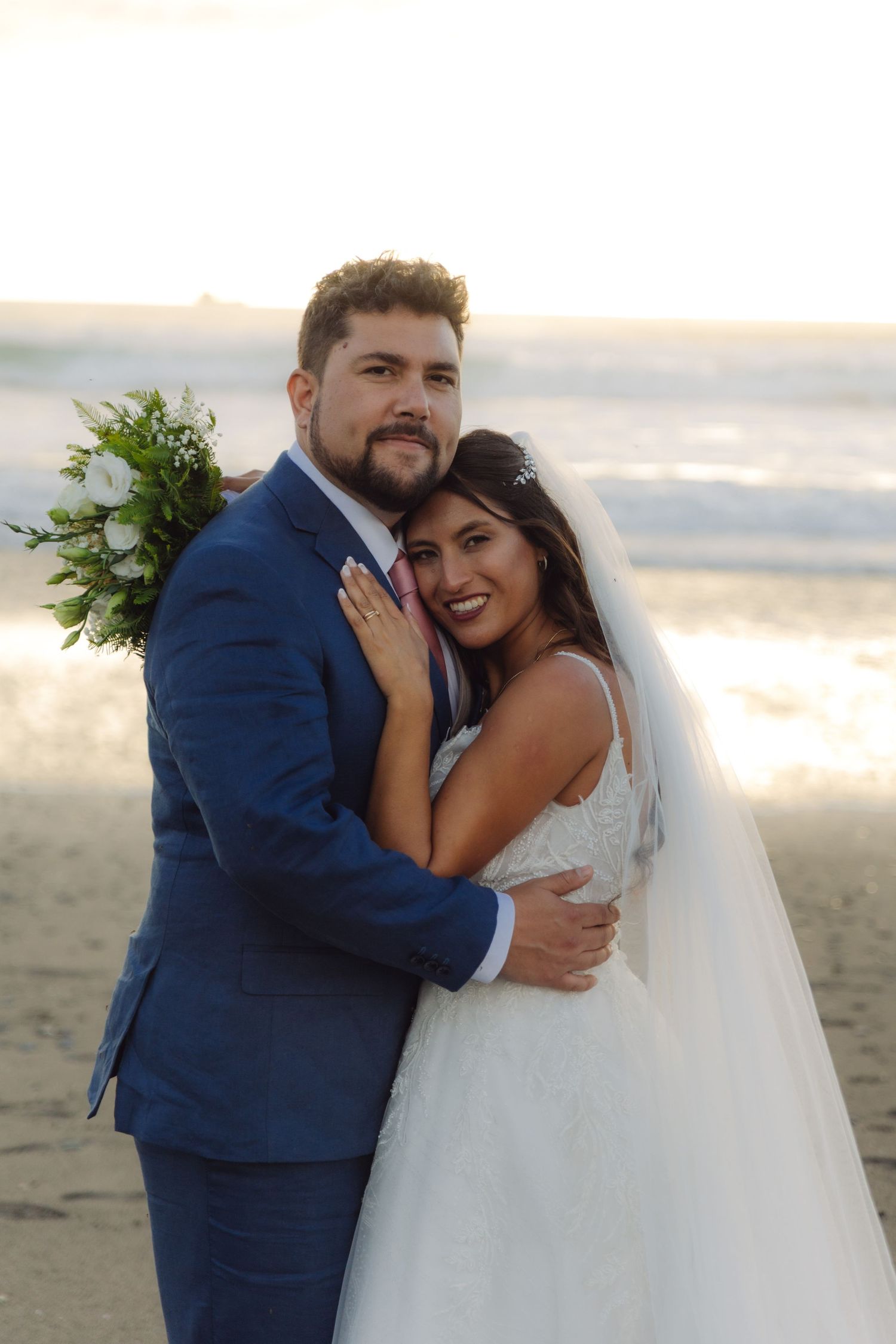 Smiling newlyweds embrace on the beach at sunset, bride in lace gown with veil, groom in navy suit.