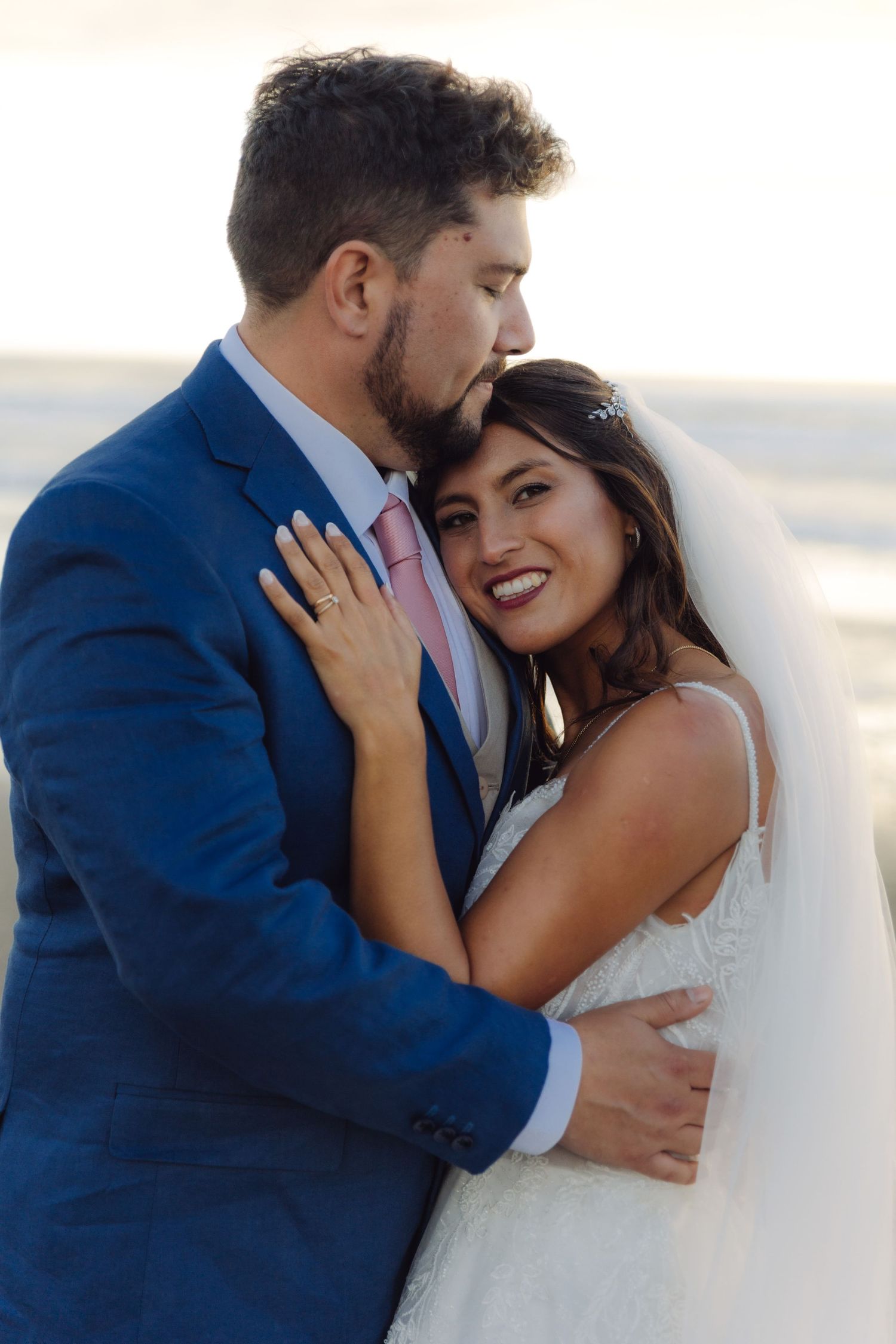 Groom tenderly kisses bride's head as she smiles radiantly, both embracing on a sunlit beach.