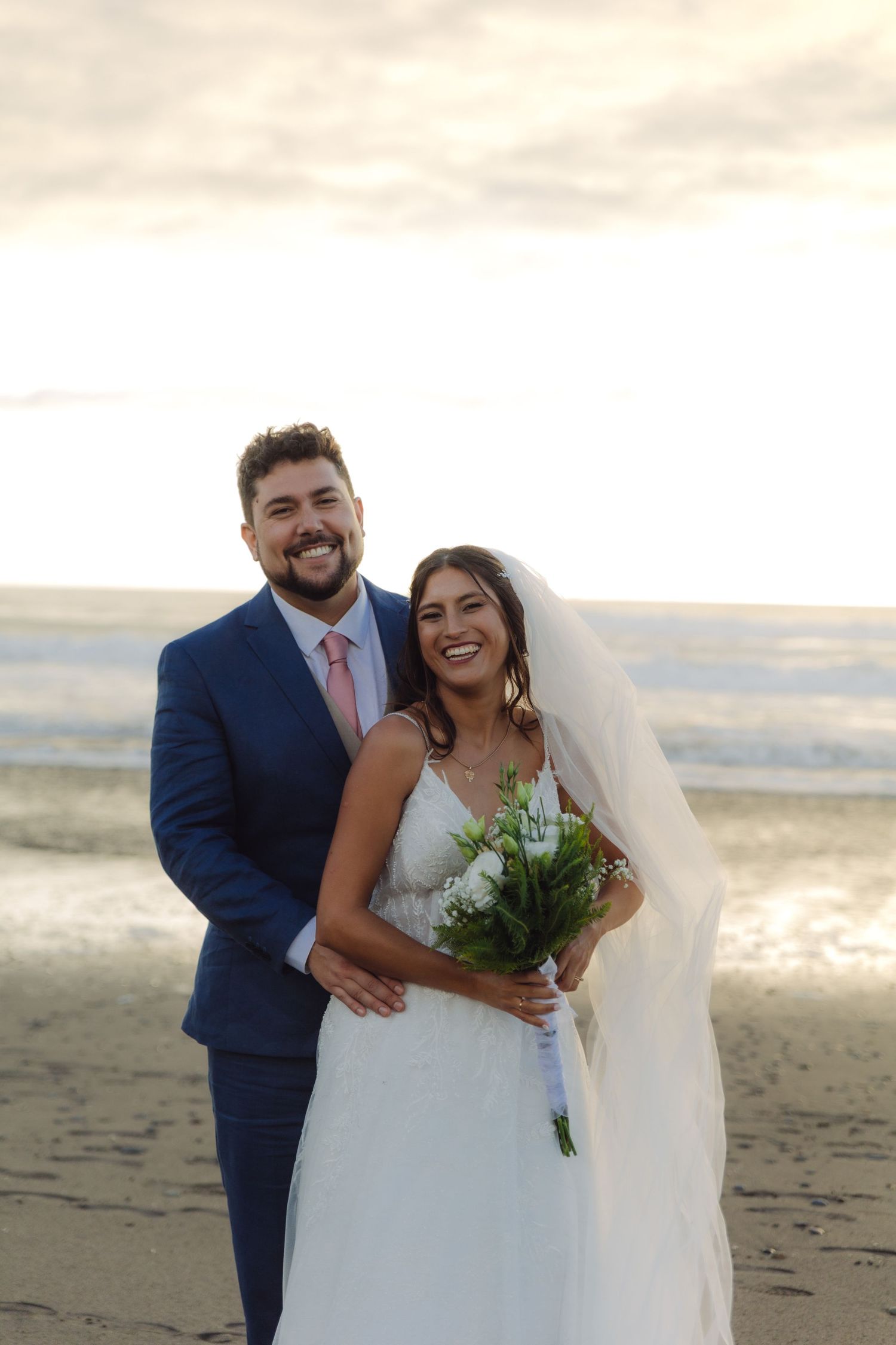 Happy bride and groom laugh together on the beach, she holding a white bouquet, ocean waves behind them.