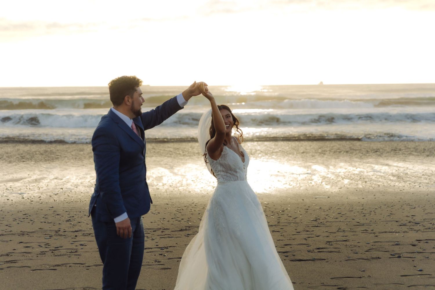 Bride and groom dancing on a sandy beach at sunset, groom in blue suit twirling bride in white gown.