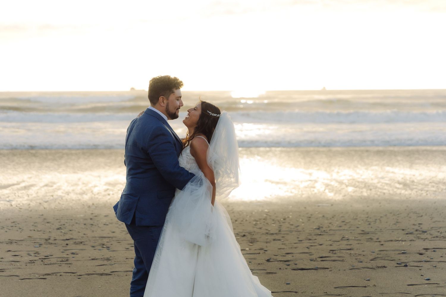 Newlywed couple sharing a romantic moment on the beach at golden hour, bride in white veil and gown.