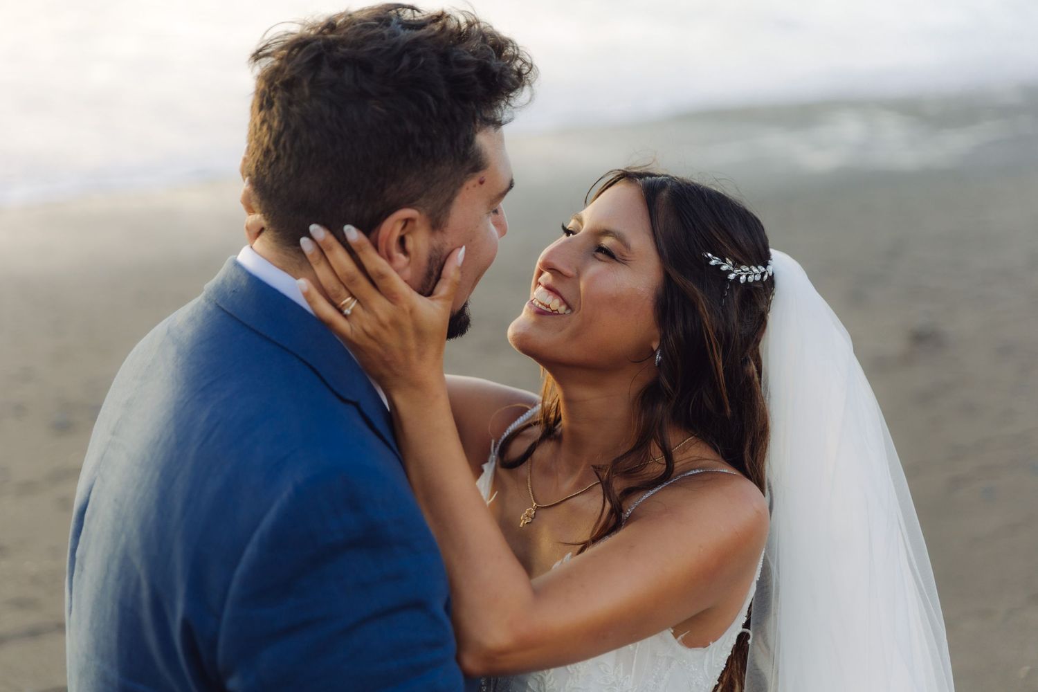 Close-up of smiling bride in floral veil gazing at groom tenderly on a beach wedding photo shoot.