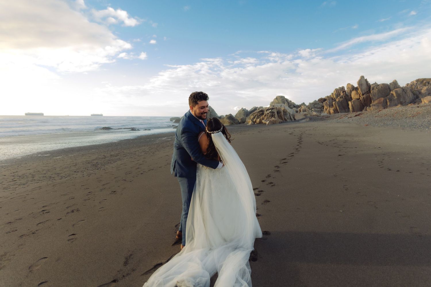 Bride and groom embracing on a scenic beach with rocky coastline, bride's long white train flowing on sand.