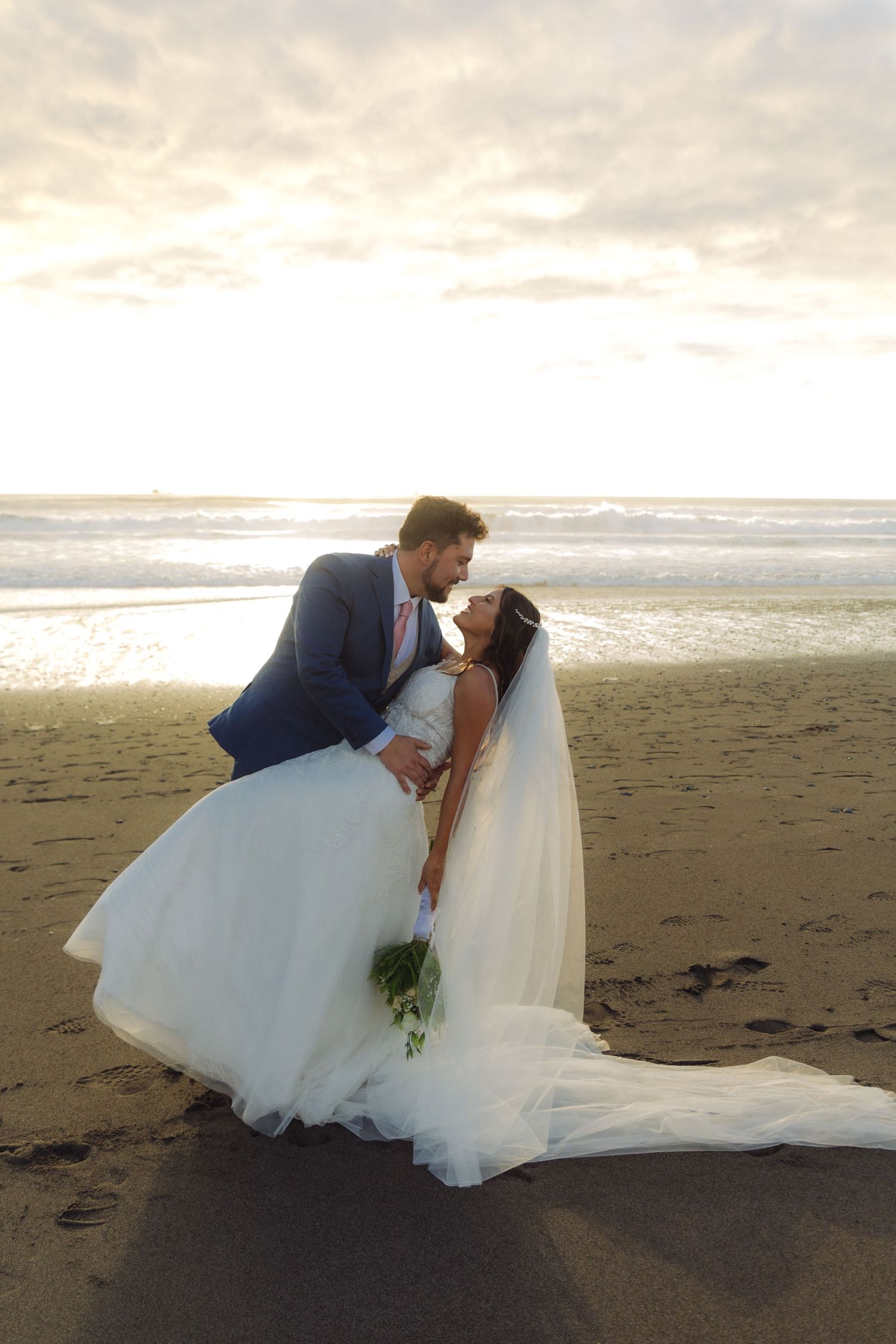 Bride and groom sharing a kiss at sunset on the beach, bride holding bouquet in flowing white gown.