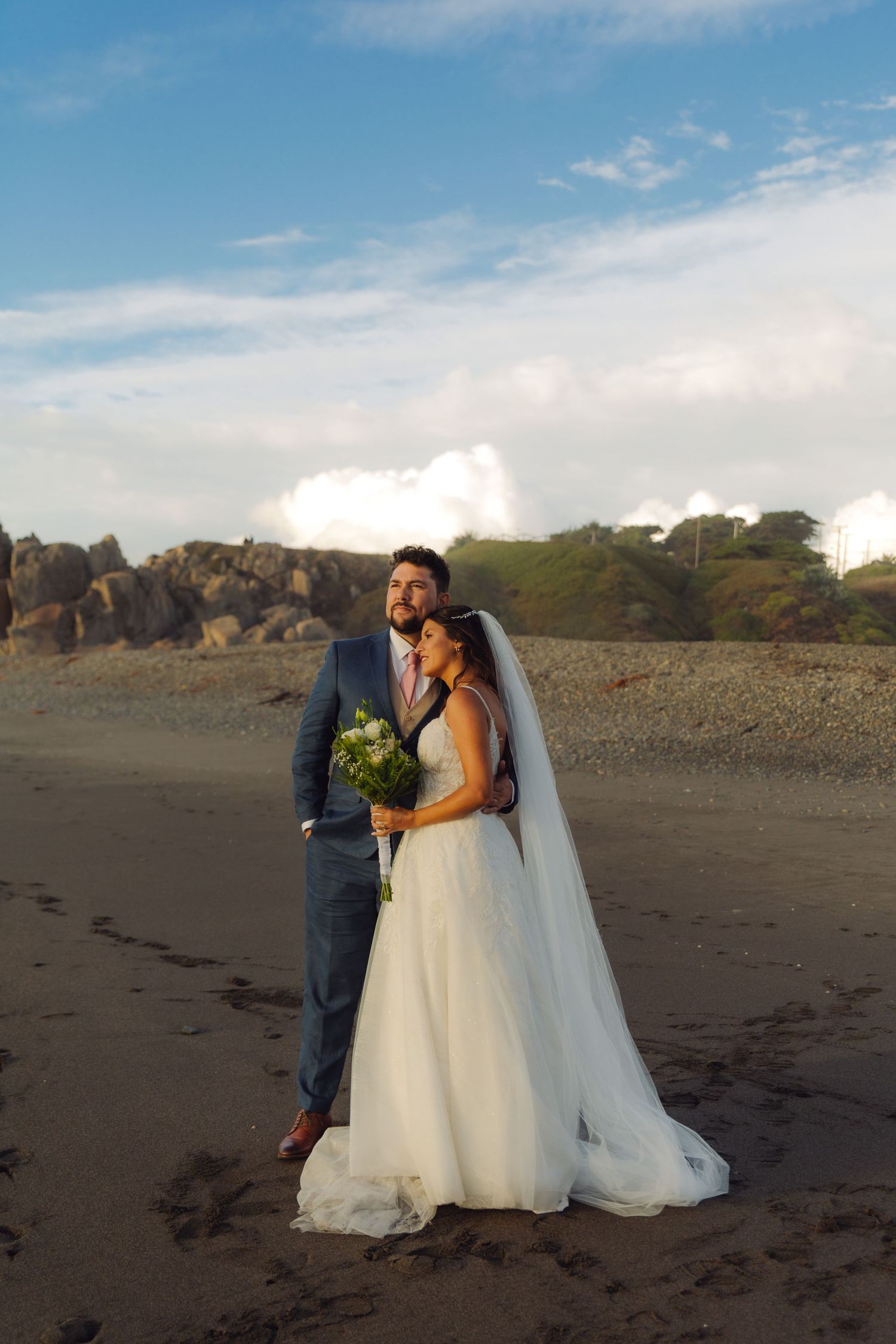 Newlywed couple posing on a sandy beach with rocky cliffs, bride holding bouquet in elegant white dress.