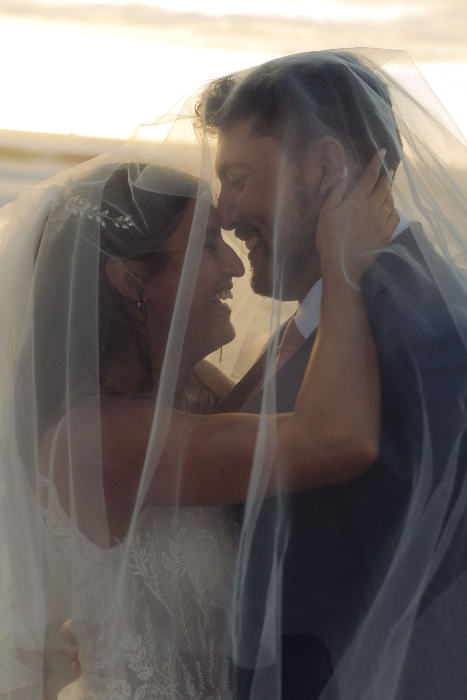 Romantic bride and groom nearly kissing under a sheer wedding veil in warm golden sunset light.
