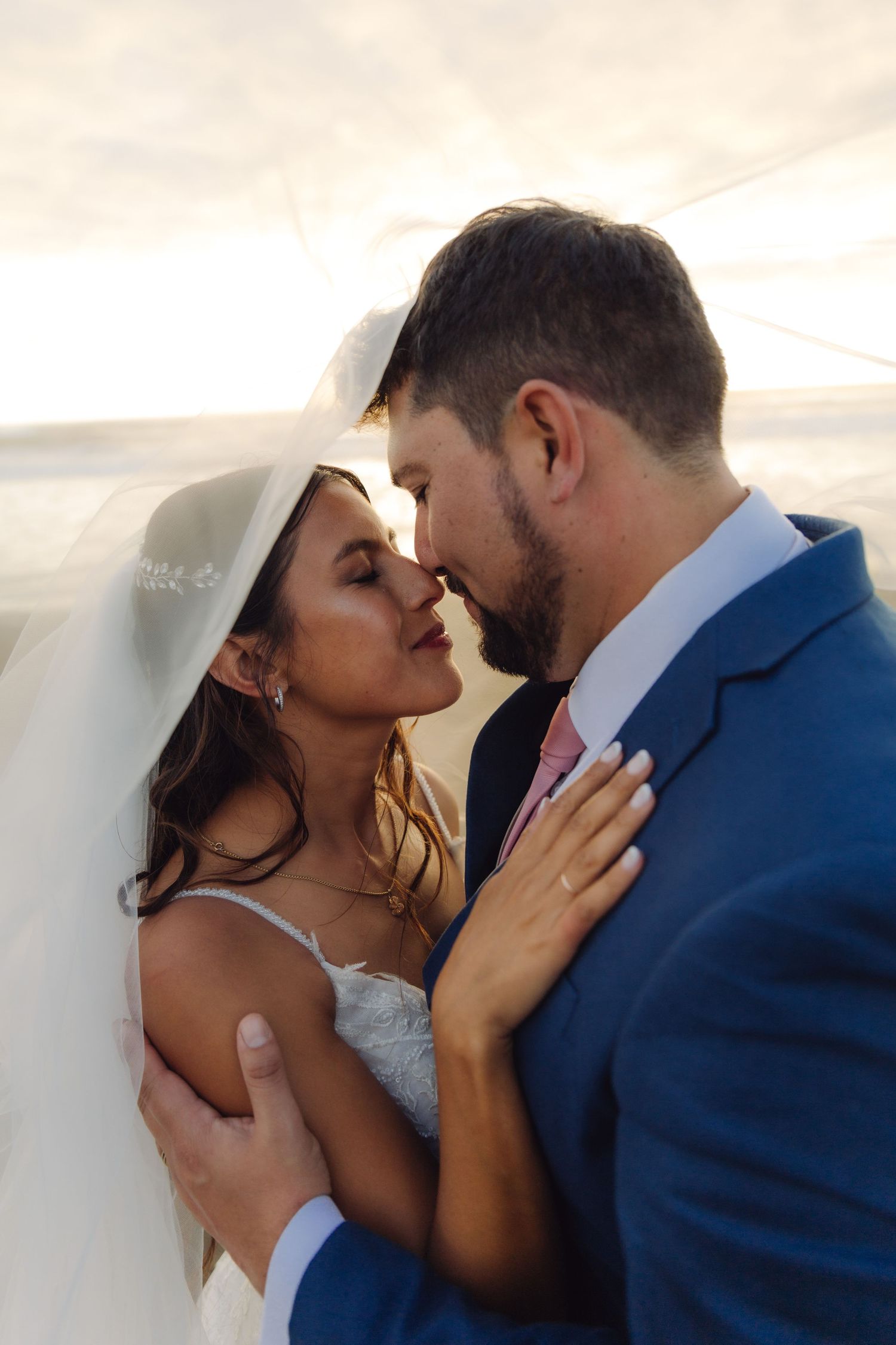 Intimate close-up of bride and groom almost kissing under wedding veil, groom in blue suit on beach.