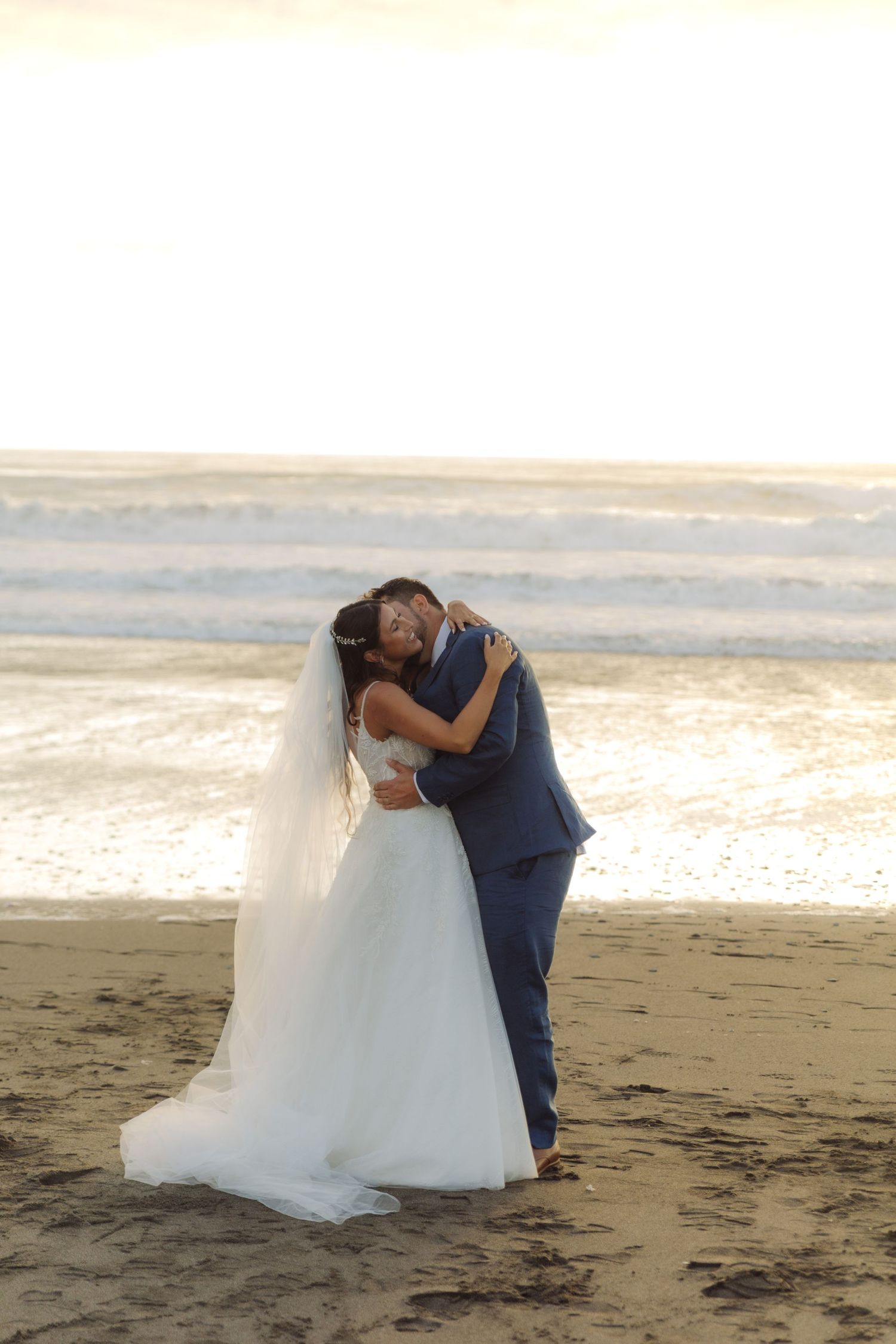 Bride and groom embracing on a sunlit beach, bride in long white gown with veil, groom in blue suit.