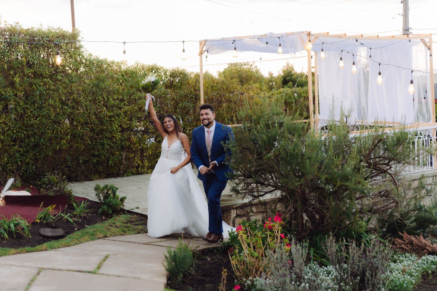 Joyful newlyweds celebrating in a garden venue with string lights, bride raising bouquet in white gown.