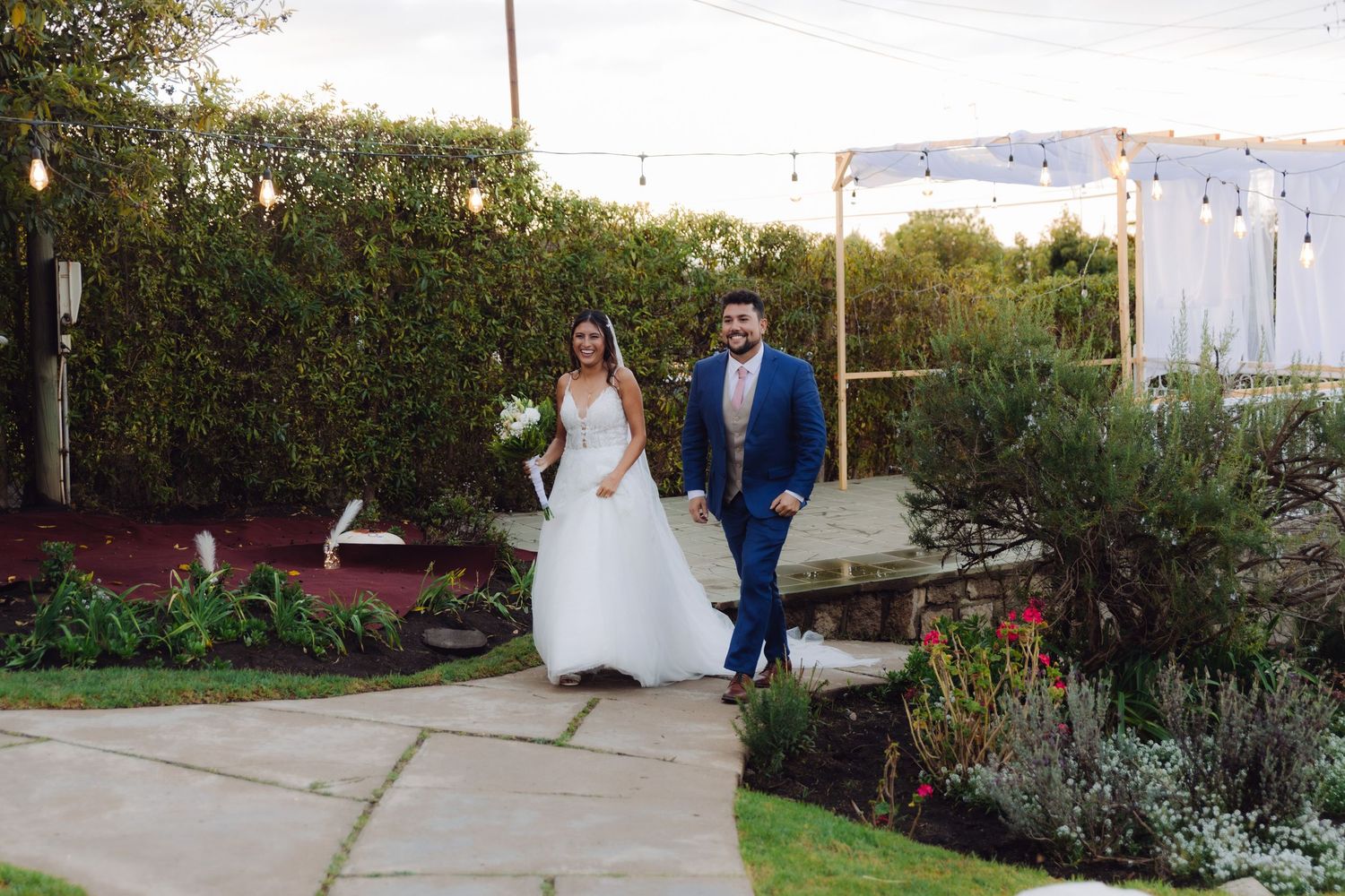 Bride and groom walking together in a garden wedding venue with string lights and lush greenery.