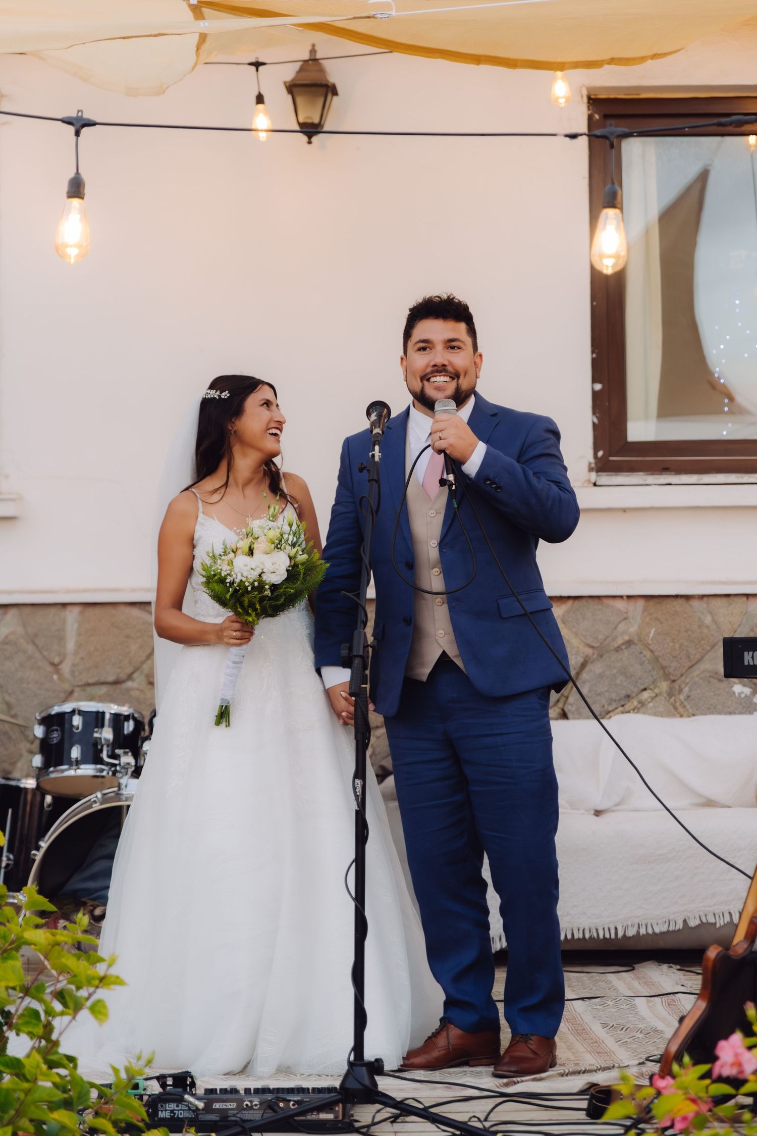 Smiling bride and groom speaking at a microphone during outdoor wedding reception with string lights.