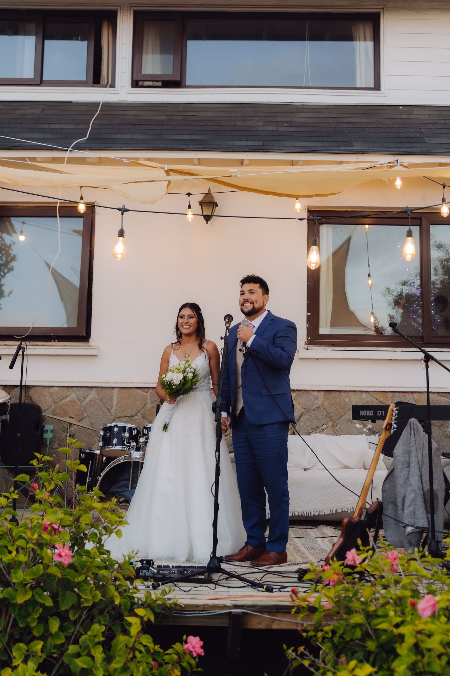 Bride holding bouquet and groom speaking at microphone on decorated outdoor wedding stage with lights.