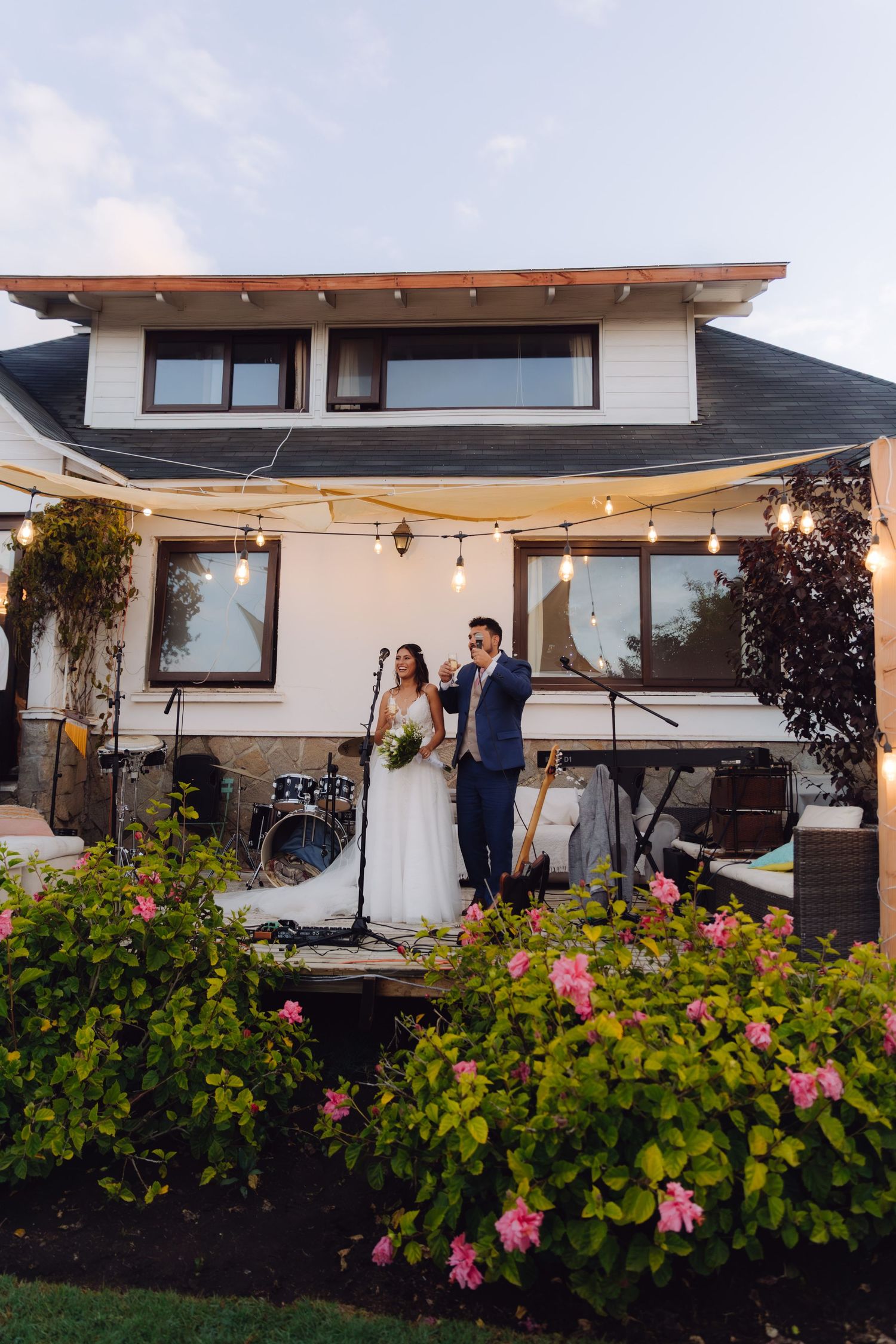 Bride and groom standing on outdoor stage at garden wedding venue with string lights and flowers.