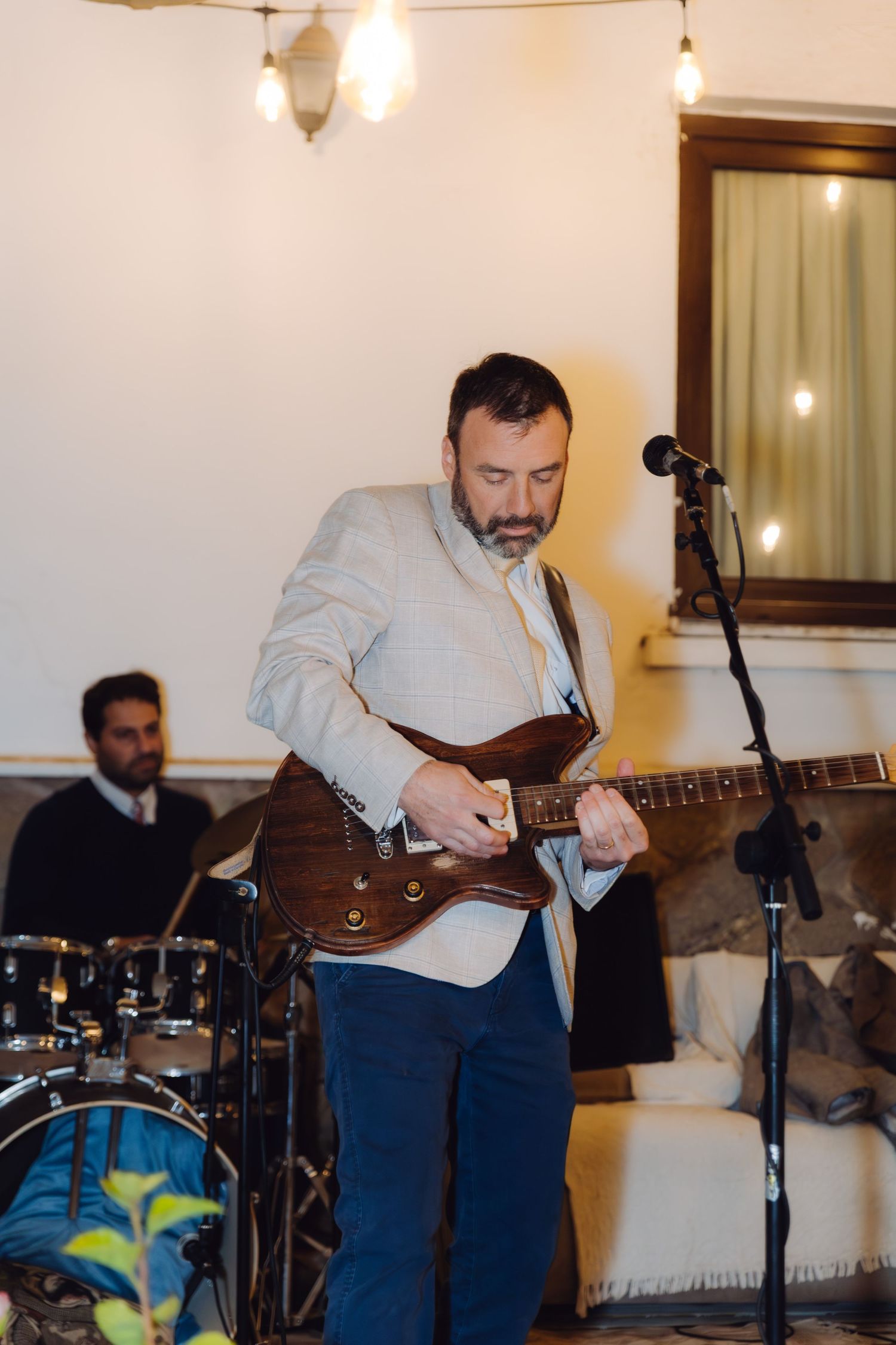Bearded musician in gray blazer playing electric guitar at indoor wedding reception with drum kit.