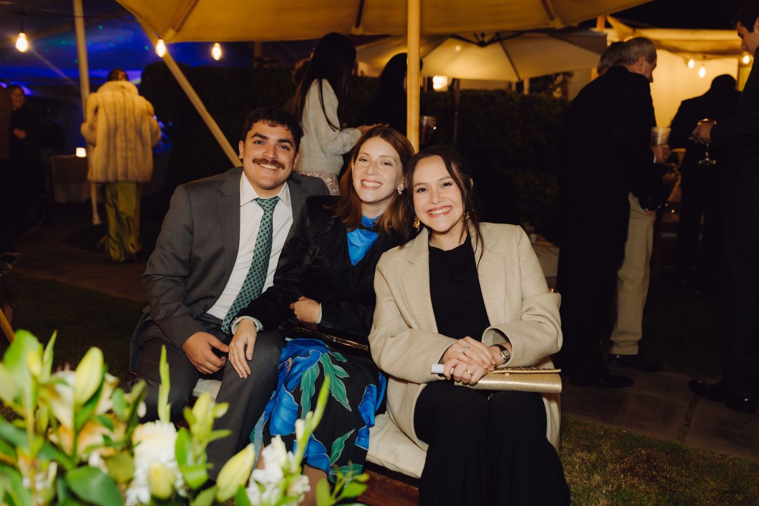 Three smiling wedding guests posing together at an outdoor evening reception under tent lights.