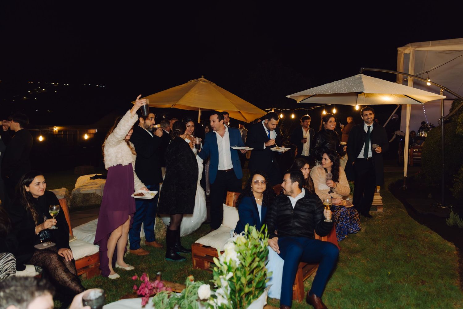 Guests mingle and dance at an outdoor evening wedding reception under yellow umbrellas with string lights.