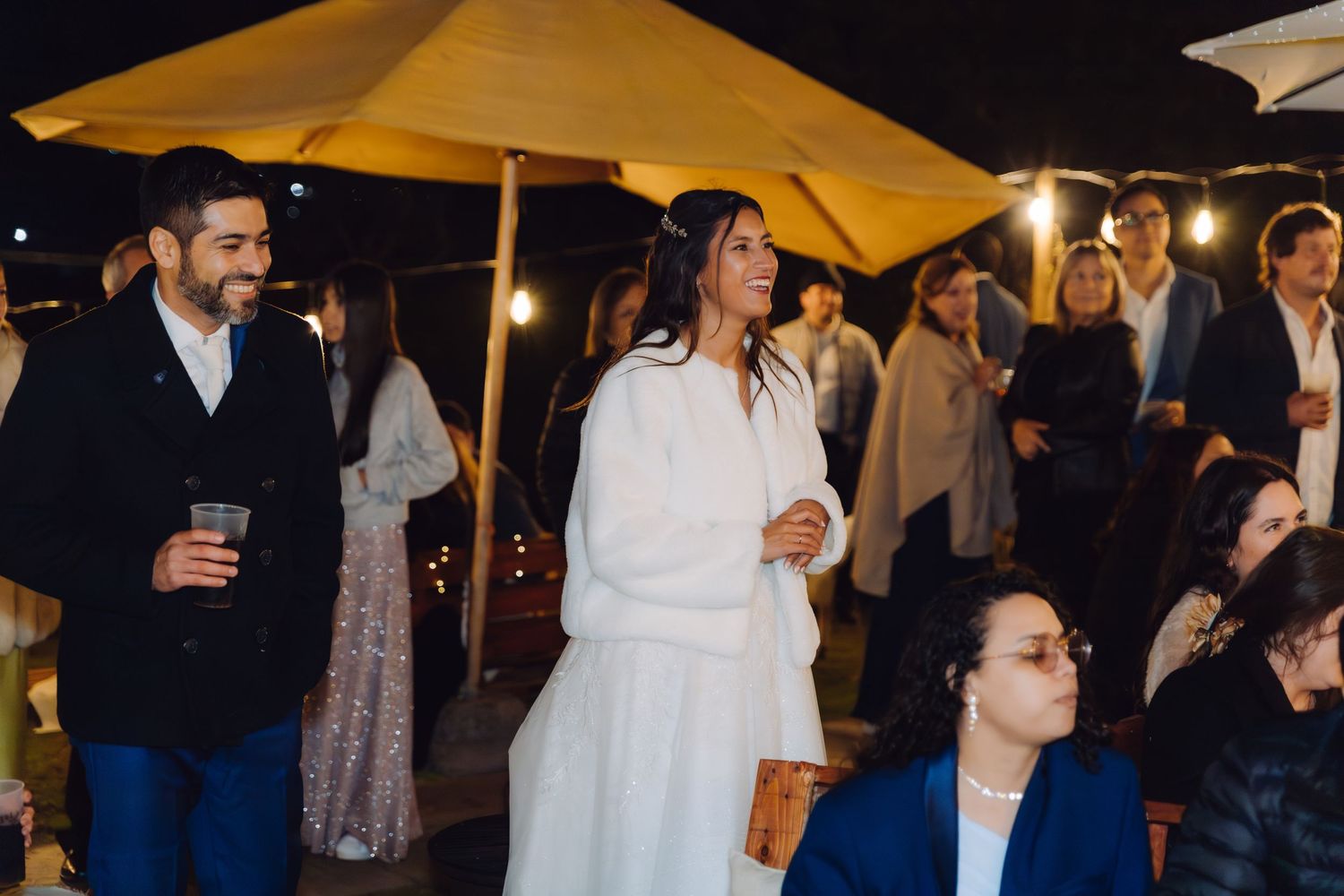 Bride in white gown stands with guests at an outdoor nighttime wedding reception under a yellow umbrella.