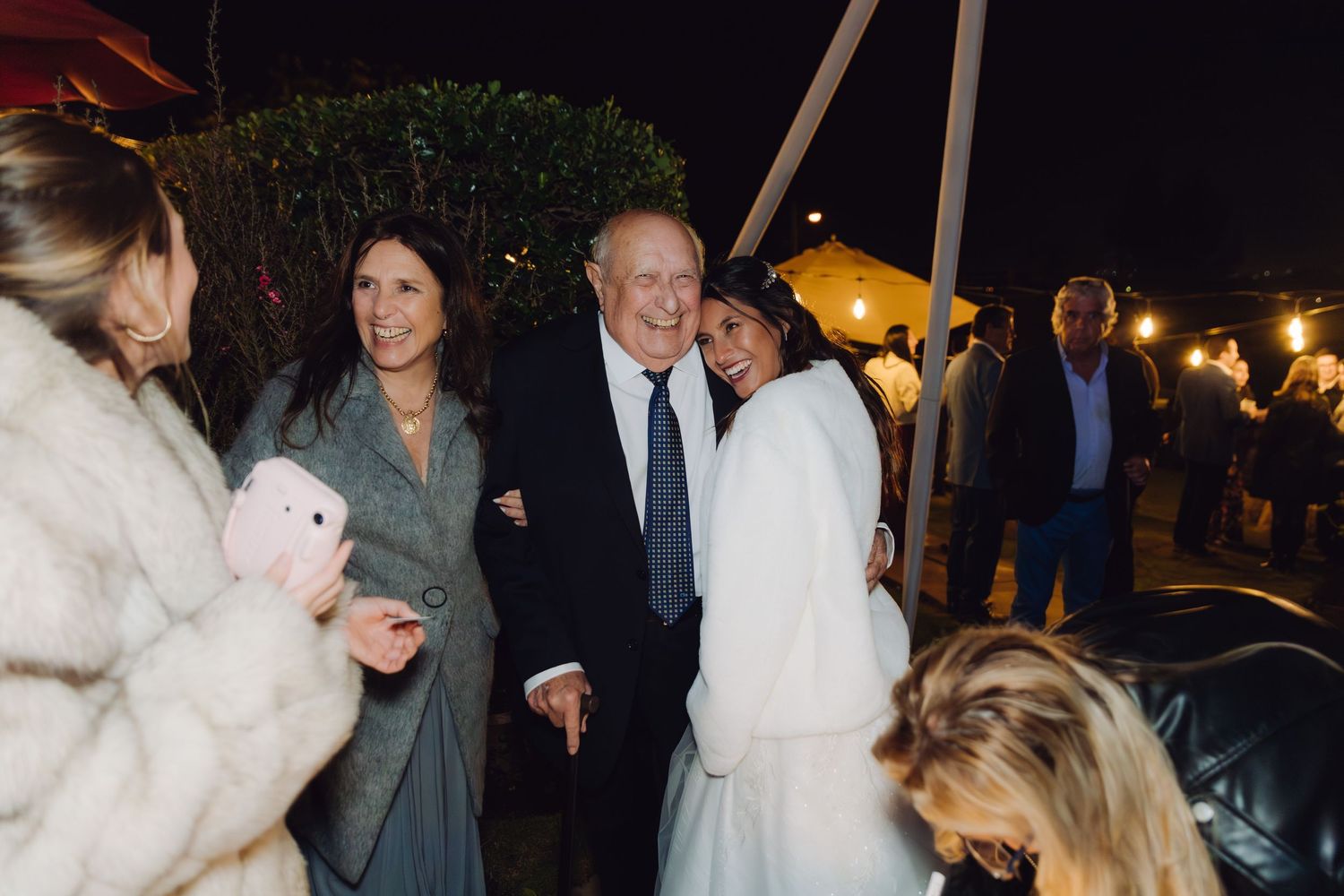 Bride in white coat smiles with an older couple at a nighttime outdoor wedding celebration.
