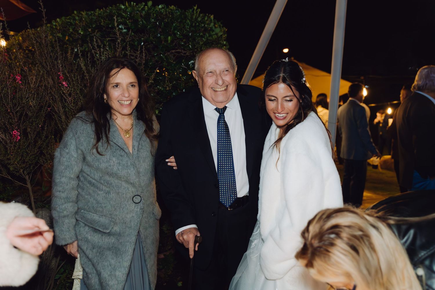 Bride in white coat poses with older couple at an elegant outdoor evening wedding reception.