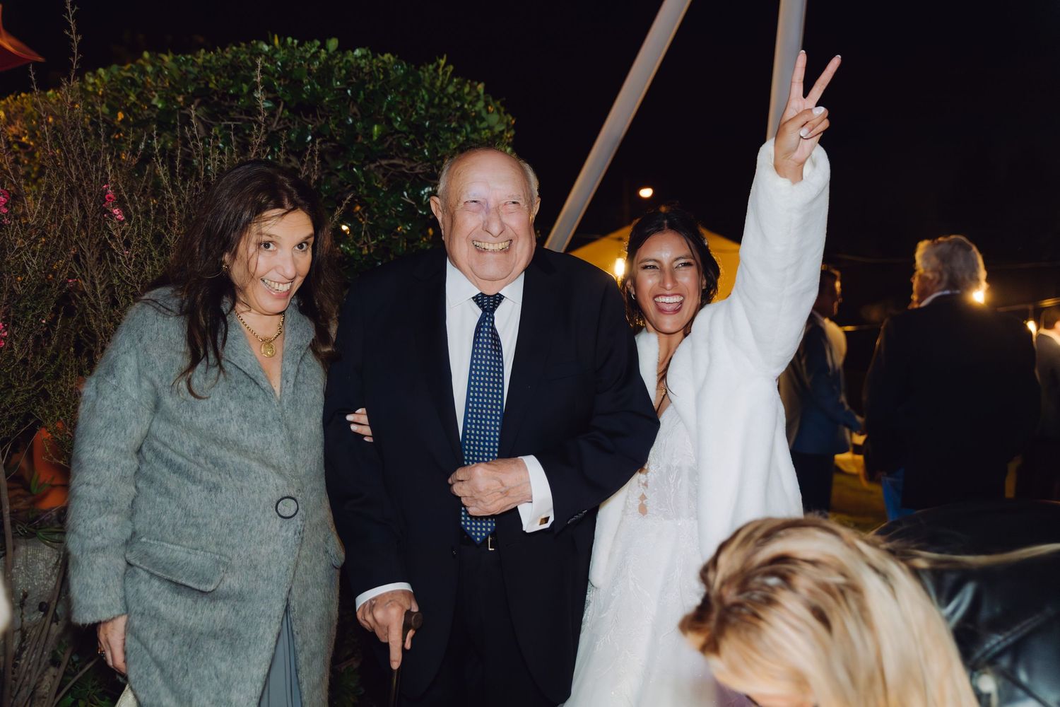 Bride in white coat gives peace sign while posing with couple at outdoor evening wedding.