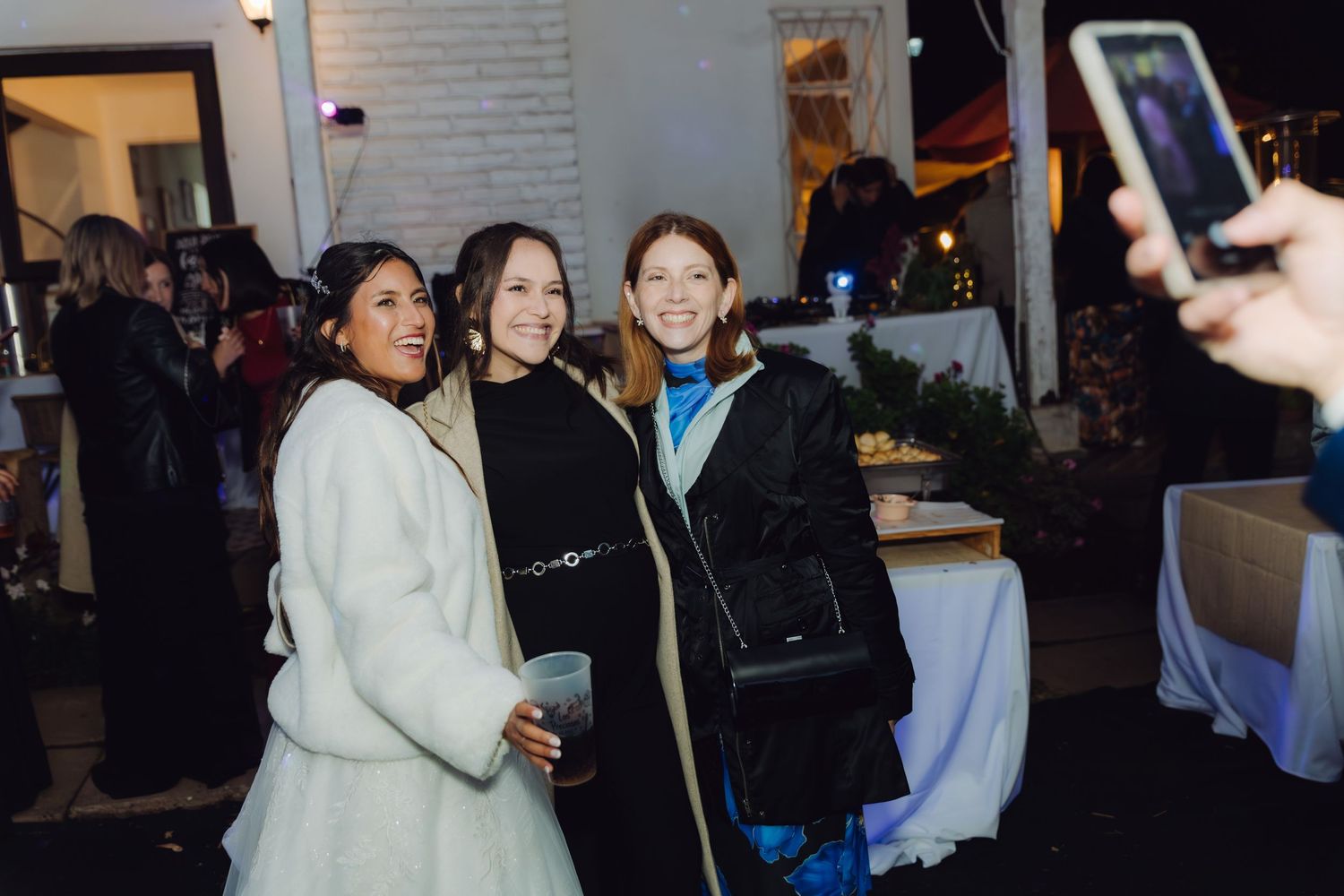 Three women smile for a photo at an outdoor evening wedding reception near a food table.