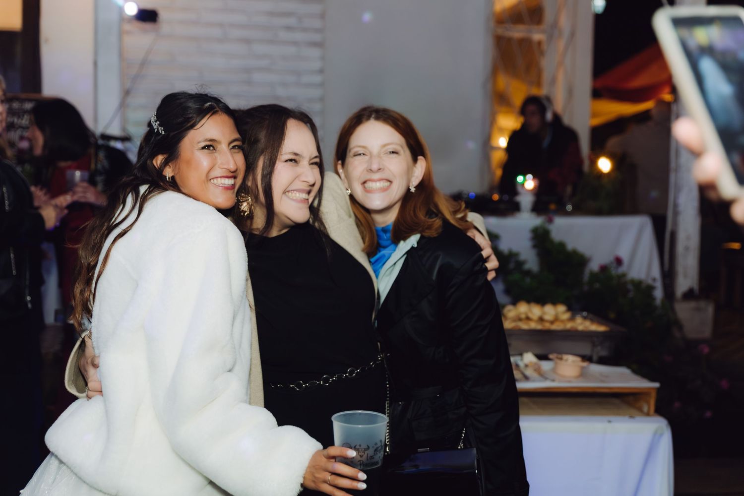 Bride in white fur coat poses with two smiling women at an outdoor wedding reception.